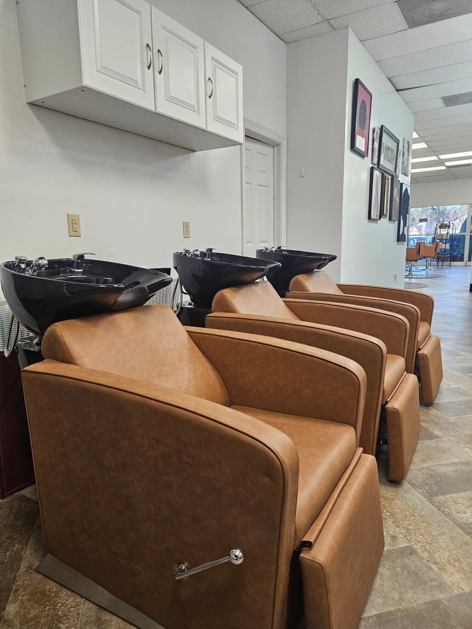 Row of brown salon chairs with black washbasins in a hair salon, white cabinets on the wall, and a window in the background.