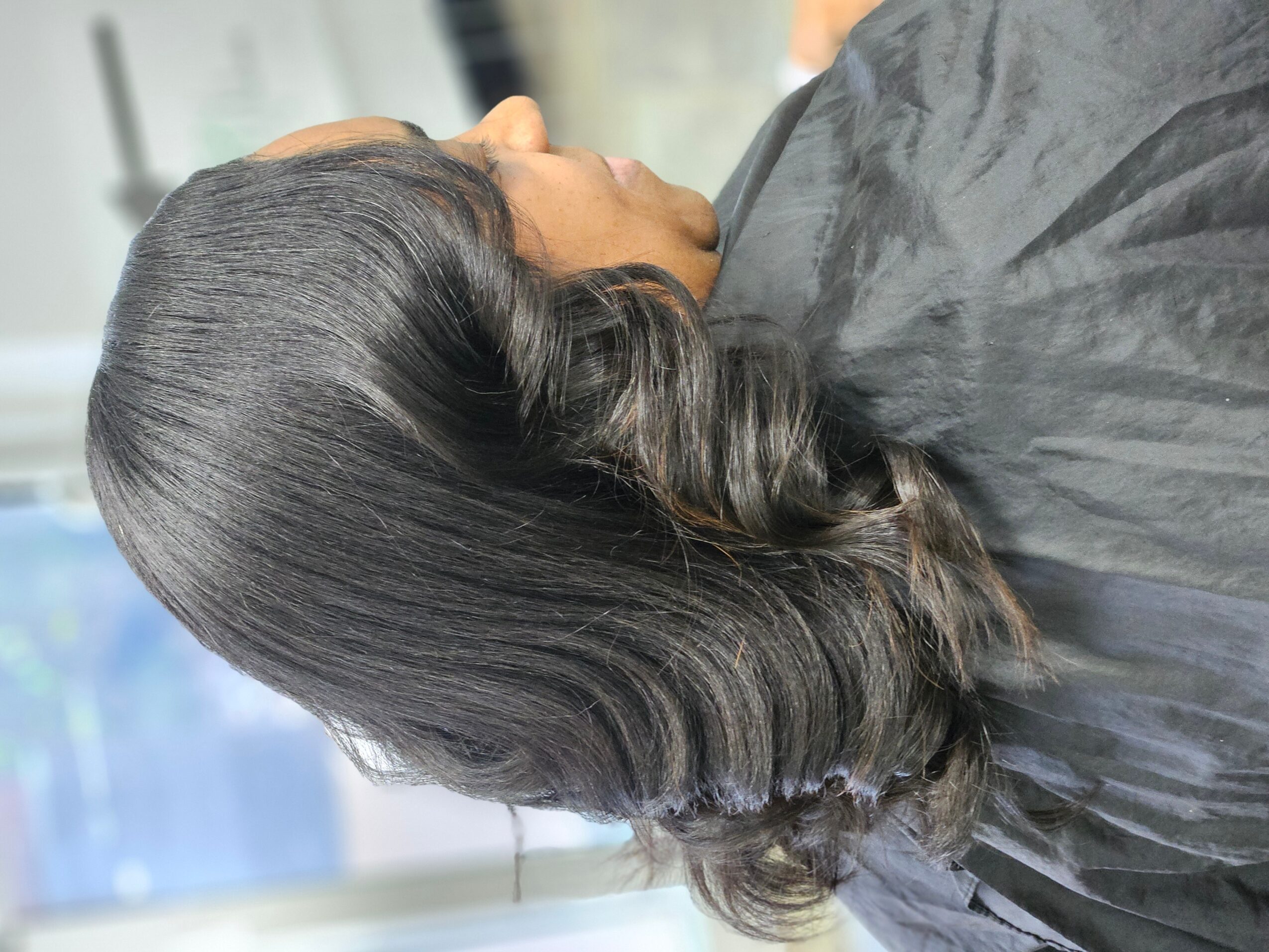 Side view of a woman with shiny, wavy, shoulder-length hair, wearing a black cape, in a salon setting.
