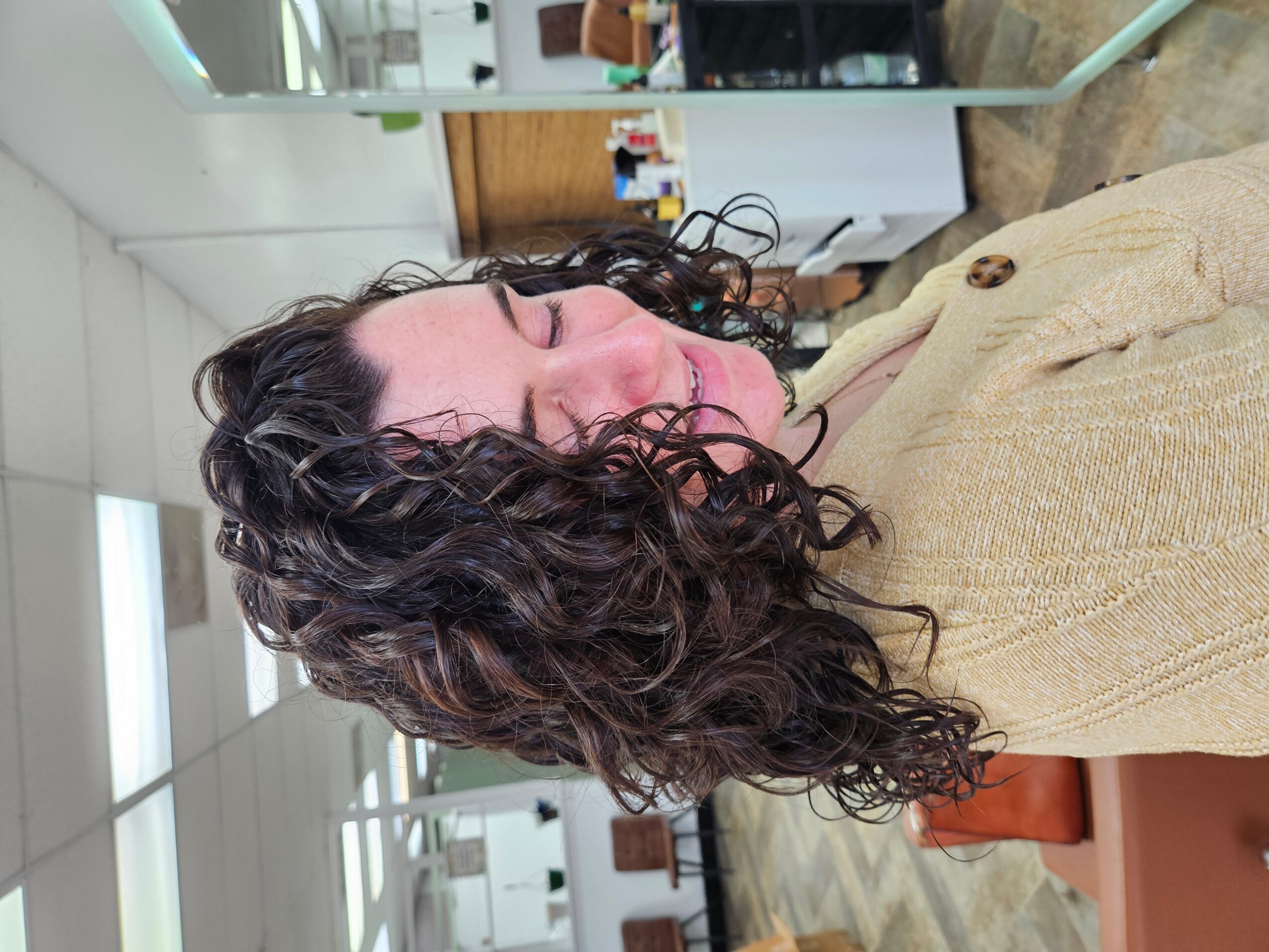 Woman with curly hair smiling, wearing a beige sweater, in an indoor setting with white walls and furniture.