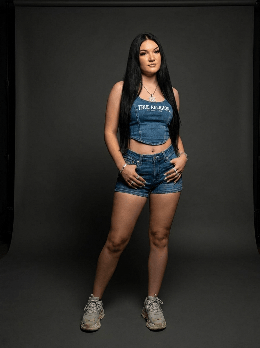 Young woman with long dark hair in denim crop top and shorts, standing with hands on hips against a black background.