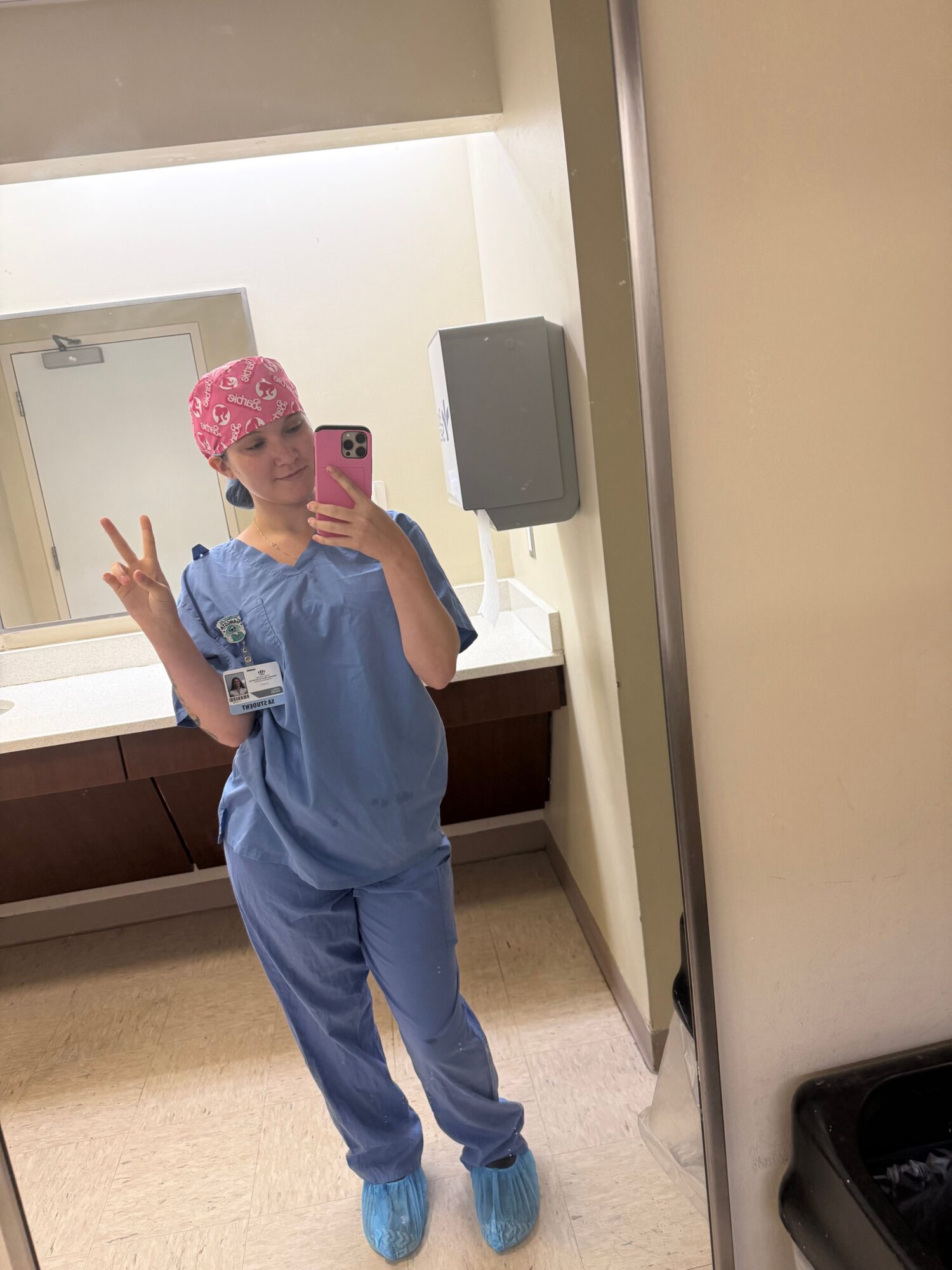 Person in blue scrubs taking a mirror selfie, making a peace sign, wearing pink cap, in a room with beige floor and white walls.