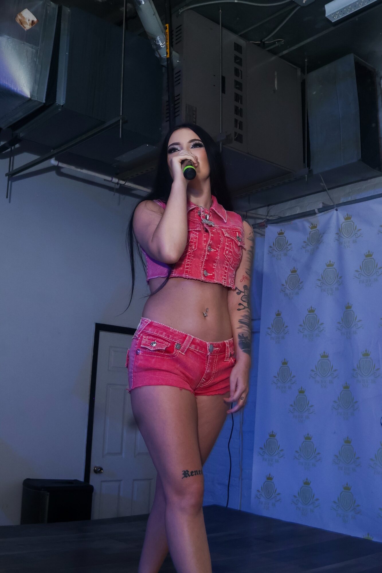 Young woman singing into a microphone on stage, wearing a pink crop top and shorts, with a backdrop and stage equipment.