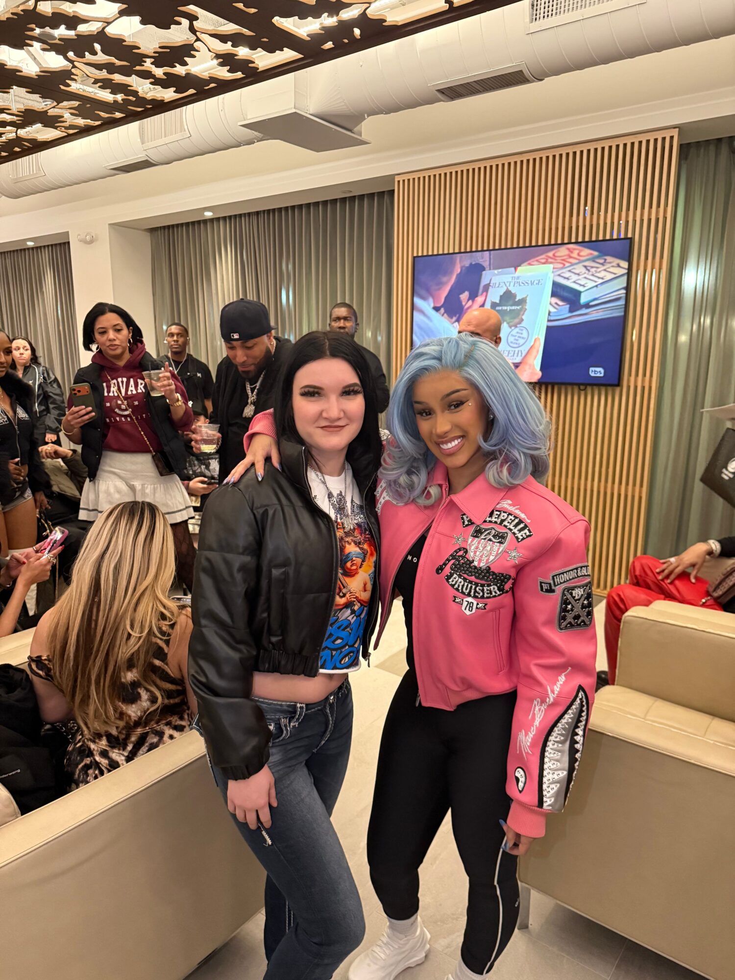Two women standing in a room with a large screen behind them, one wearing a pink jacket and the other a black jacket, with several people in the background.