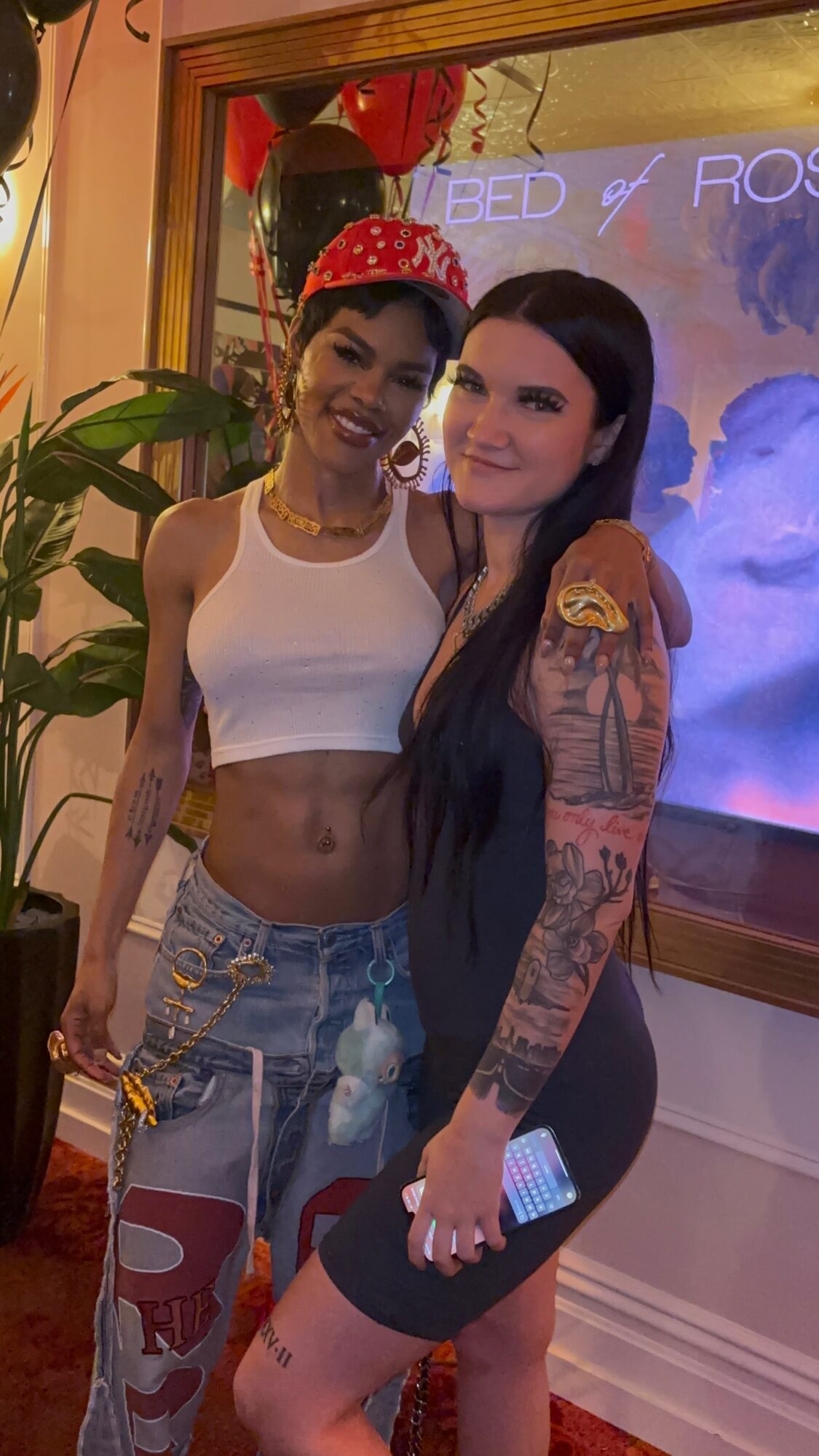 Two women smiling indoors, one wearing a white crop top and denim shorts, the other in a black dress with tattoos, holding a phone.
