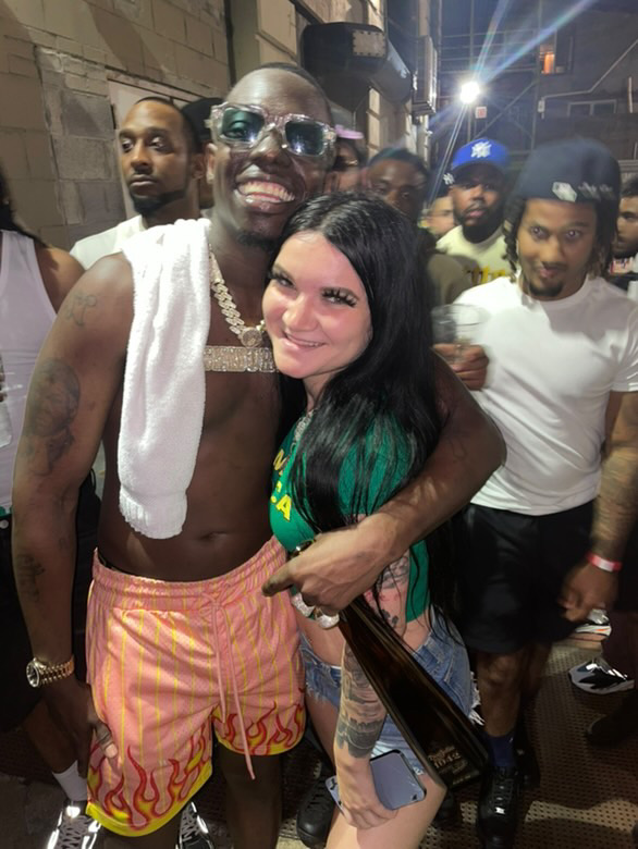 Group of people, with a shirtless man wearing sunglasses and a towel, smiling and hugging a woman with long dark hair, in a lively setting.
