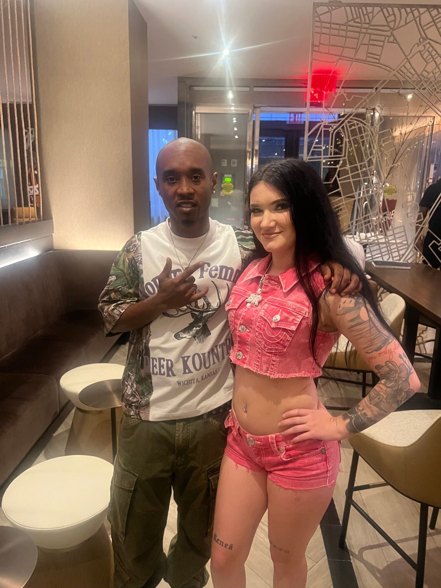 Two people standing indoors, one with tattoos and long dark hair, the other with short hair, in casual clothing.