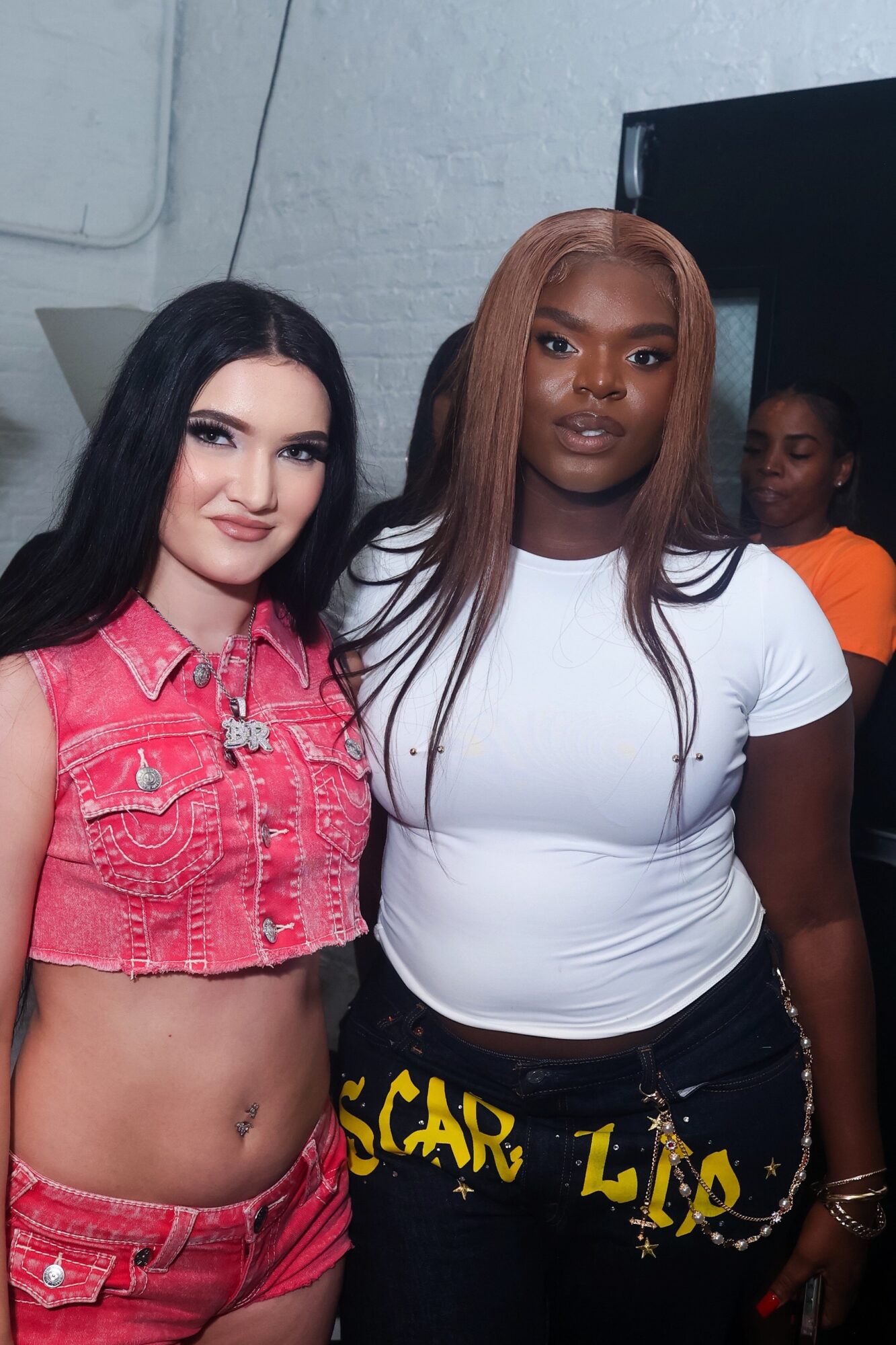 Two women standing together, one with long black hair in a pink denim crop top, the other with long brown hair in a white t-shirt, in a room with a gray wall and a black screen.
