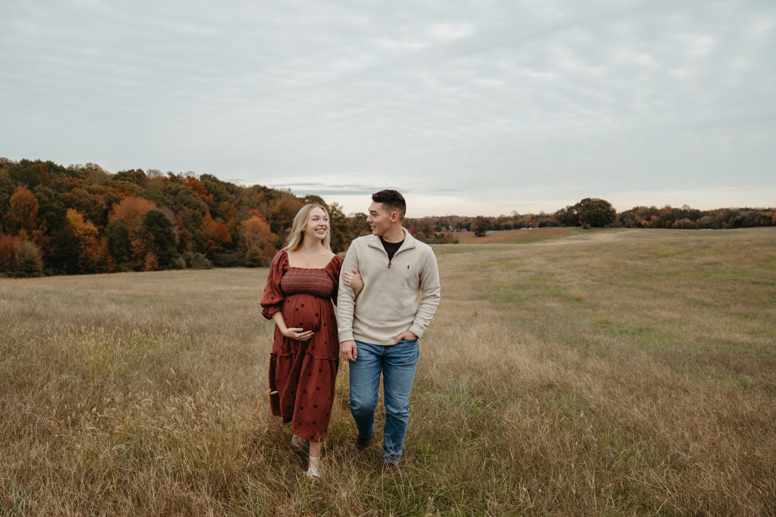 A couple walking in a grassy field with trees in the background, overcast sky, woman holding a baby bump, smiling at each other.