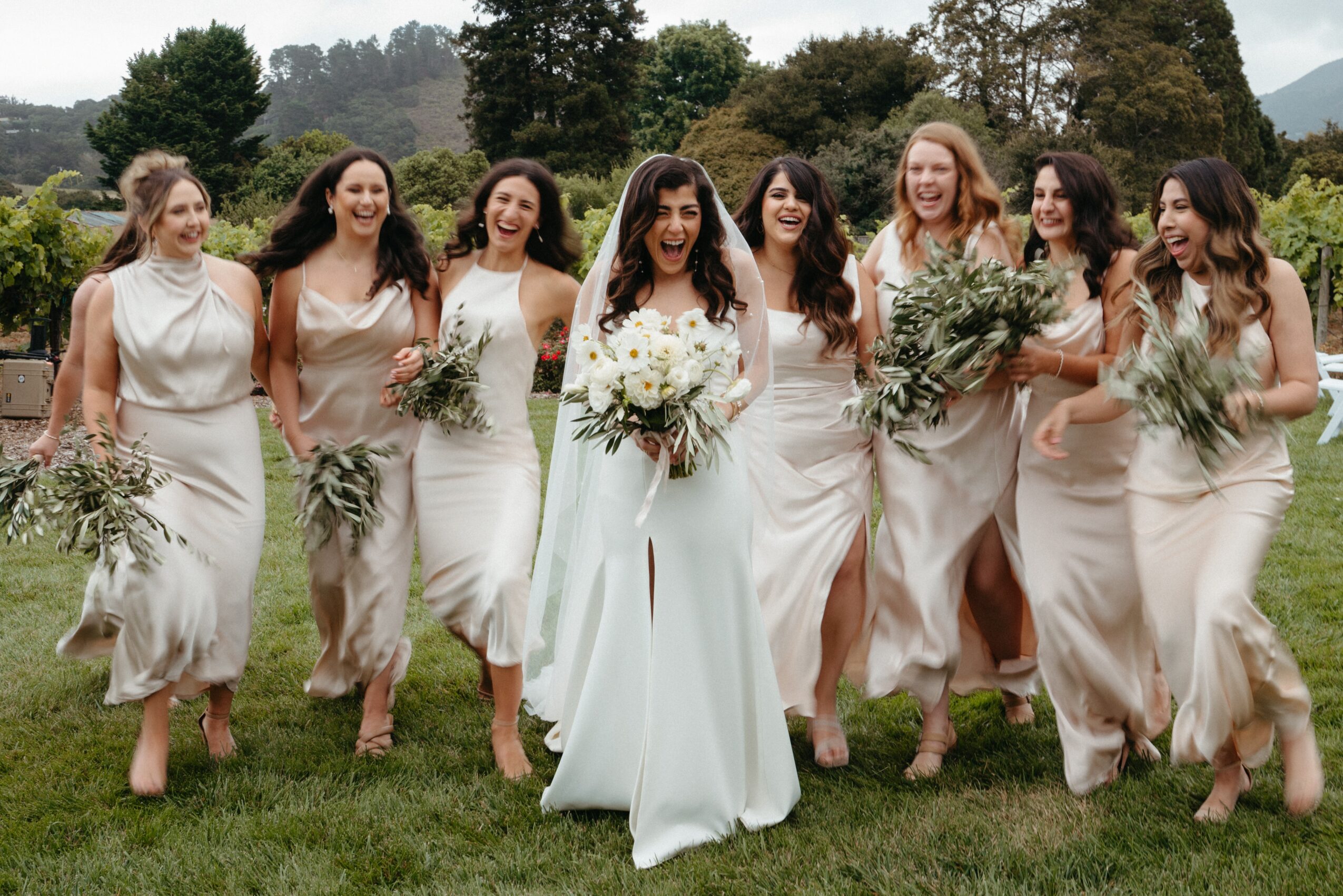 Group of women in white dresses walking on grass, holding bouquets, outdoors with trees in background.