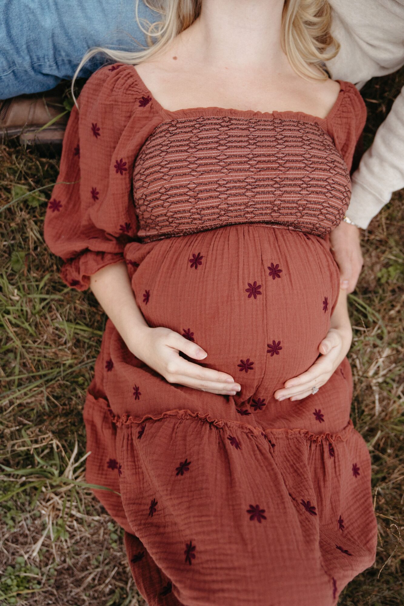 Person lying on grass, wearing a reddish-brown dress with floral patterns, holding their pregnant belly with both hands.