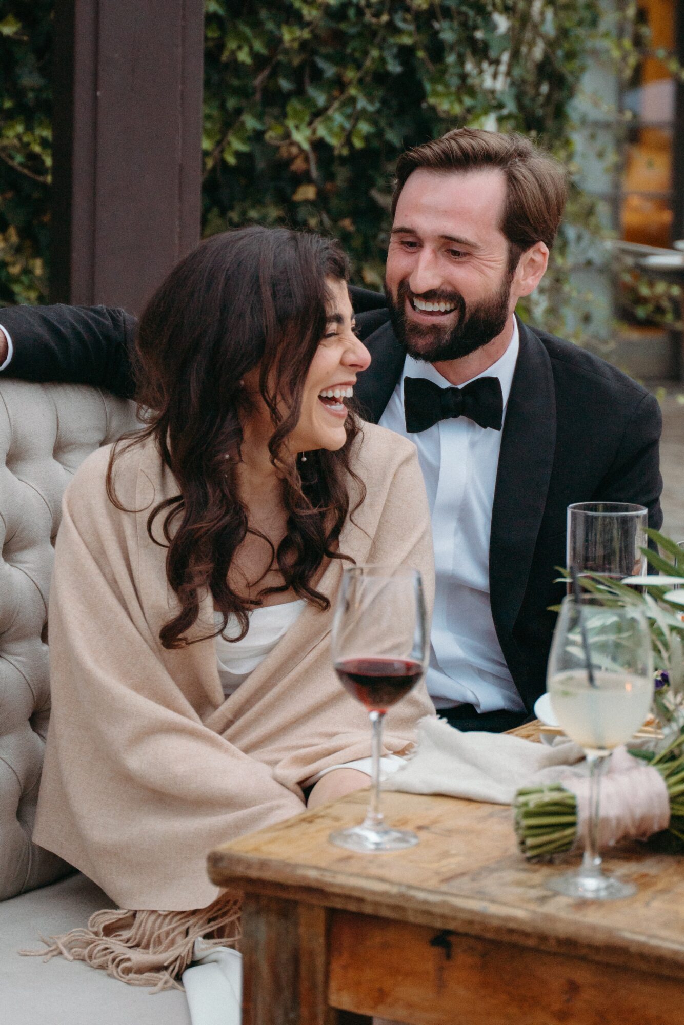 A smiling couple sitting at a table with wine glasses, outdoors with greenery in the background.