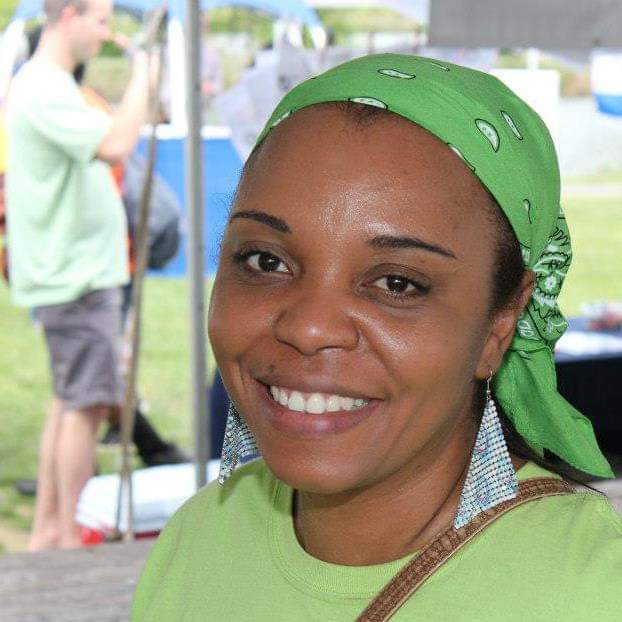 Smiling woman wearing a green bandana and shirt, outdoors with people in the background.