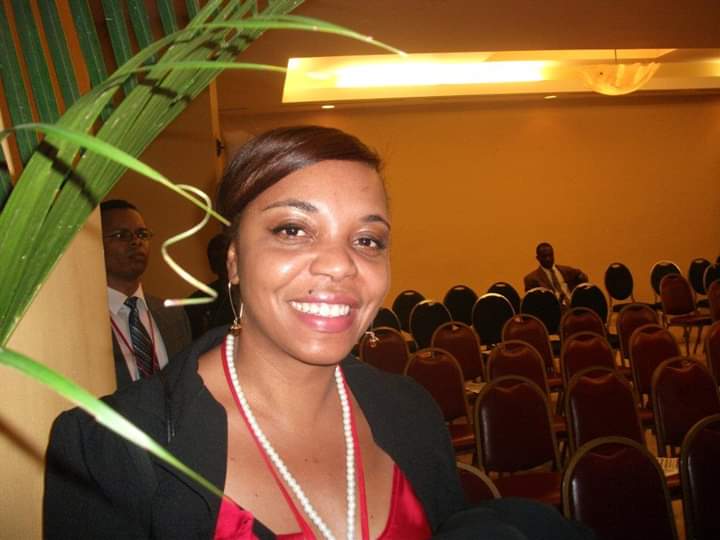 Smiling woman with short hair, wearing a black blazer, red top, and pearl necklace, in a conference room with empty chairs.