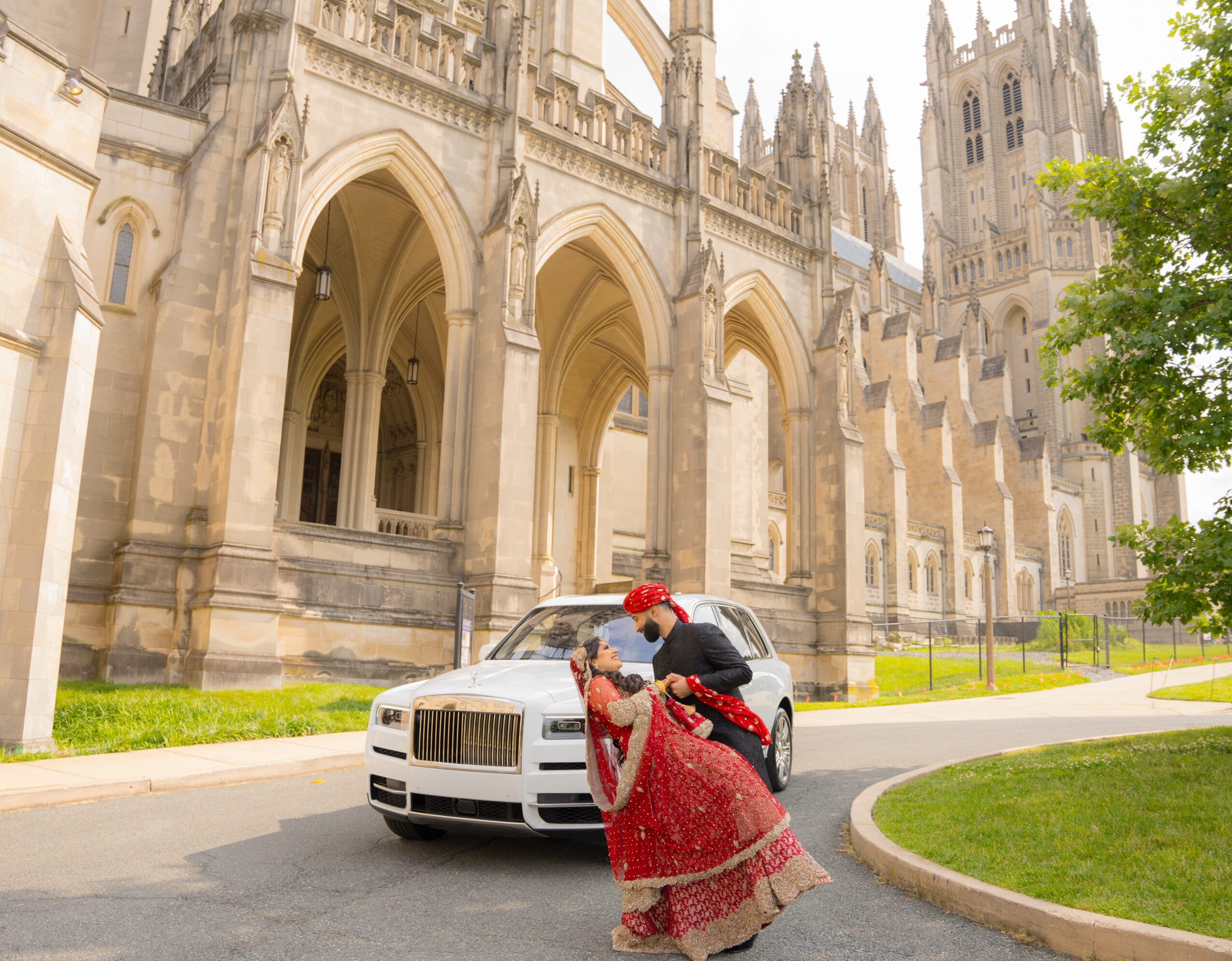 A couple in traditional attire kissing in front of a luxury car outside a cathedral, with green grass and trees nearby.