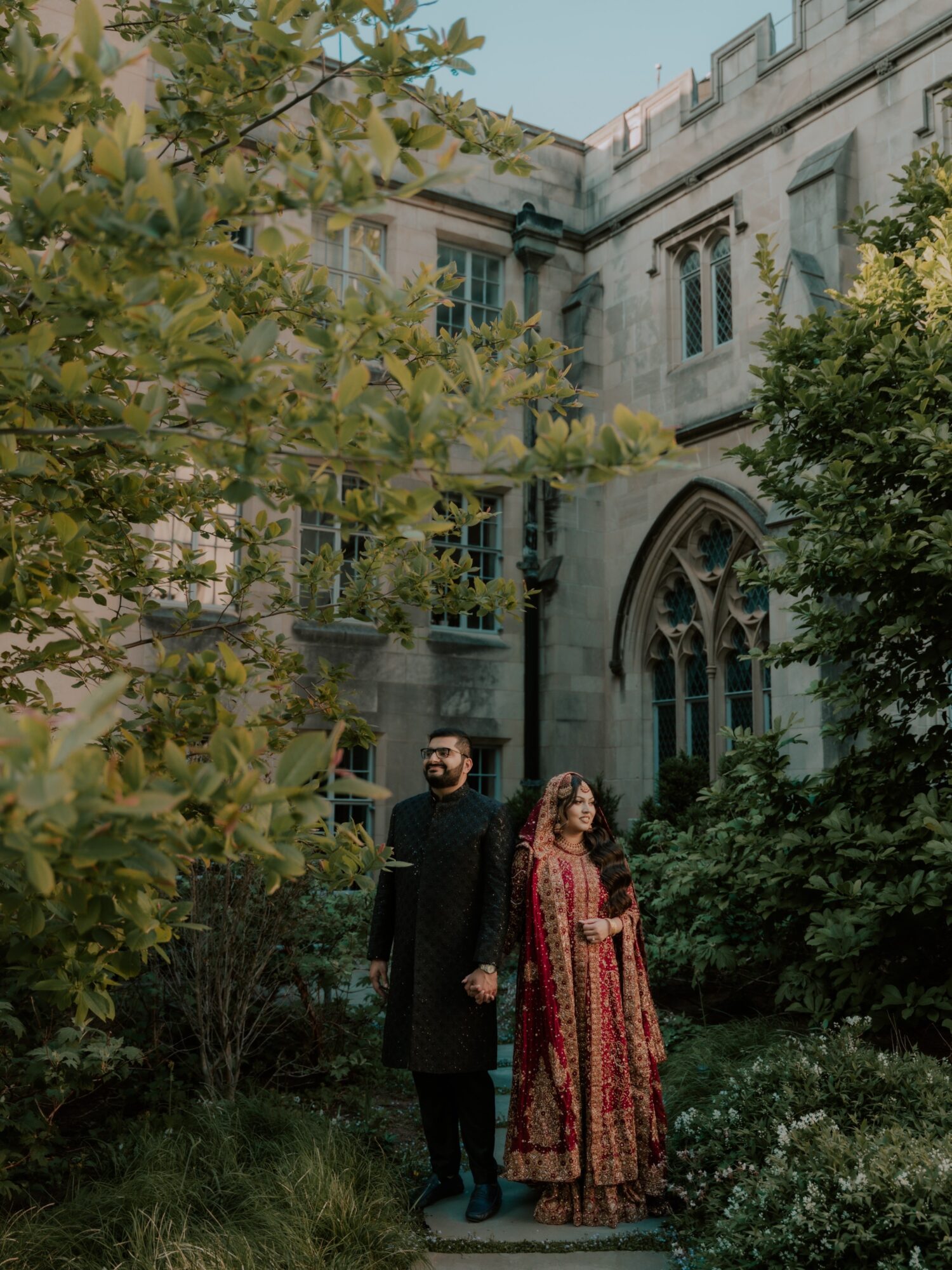 A man and woman stand outdoors in front of a historic building with arched windows, surrounded by trees and greenery.