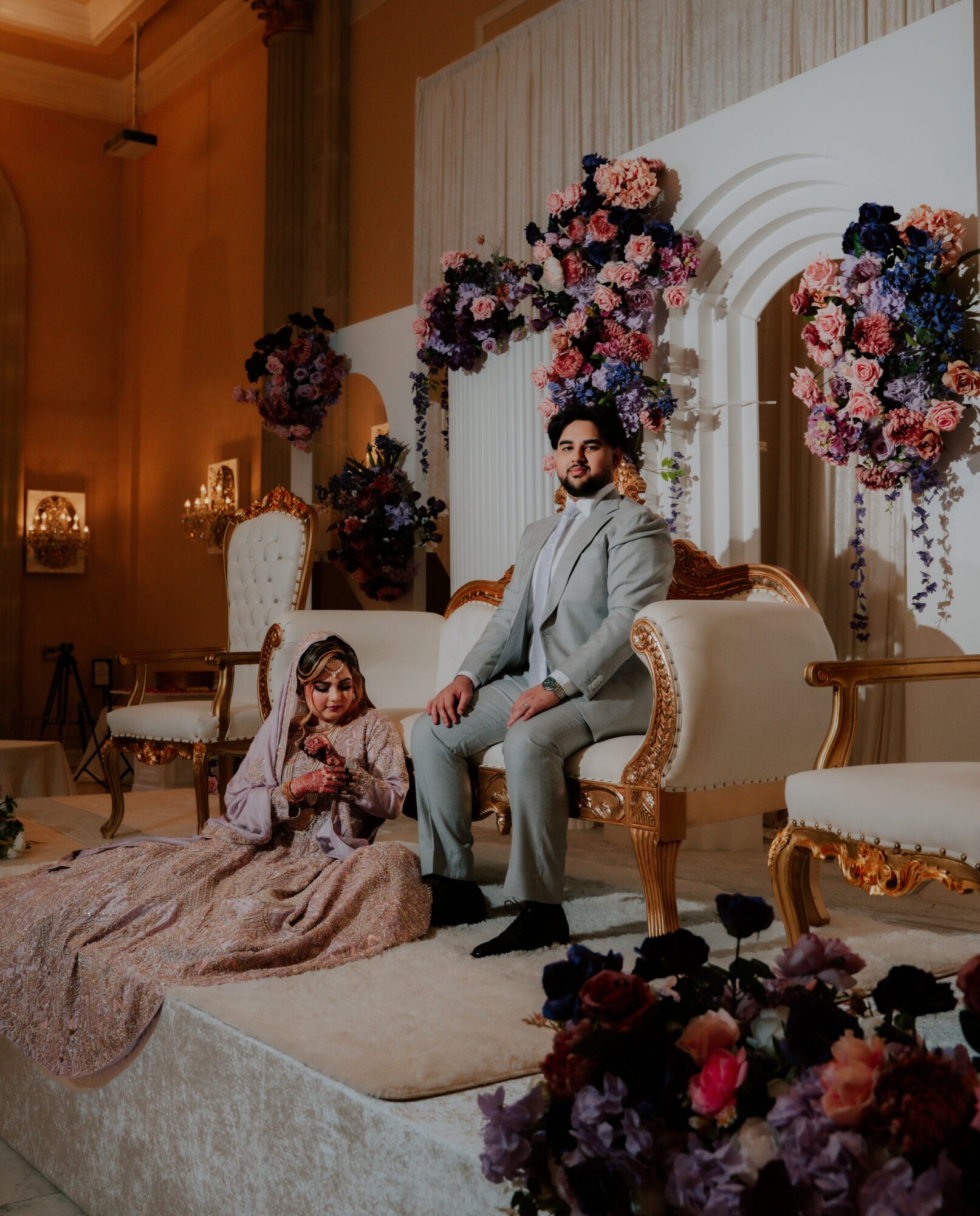 A man and a young girl sit on a decorated stage with floral arrangements and elegant furniture, in a formal setting.