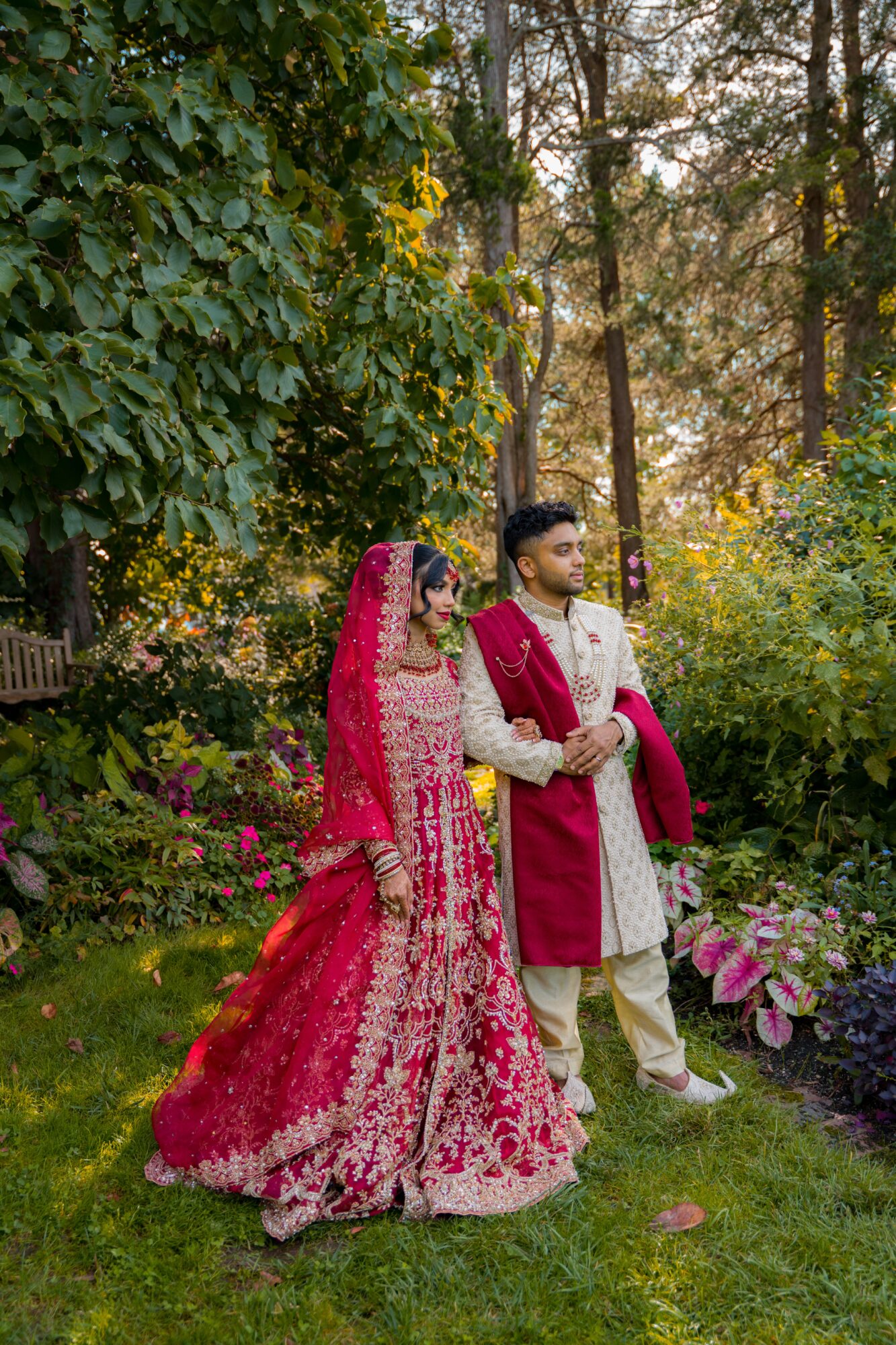 A bride and groom in traditional attire stand outdoors in a garden with trees and flowers.
