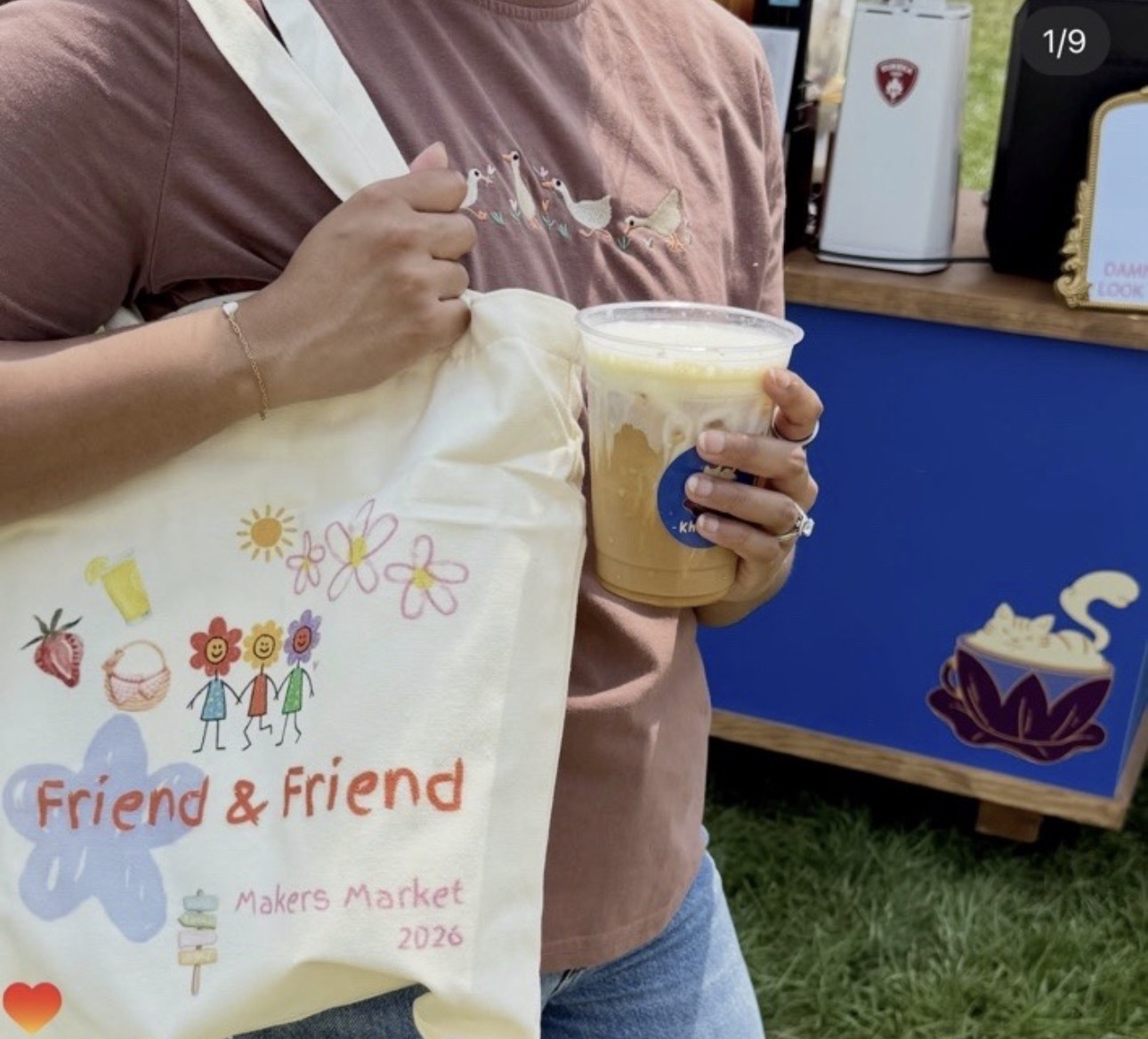 Person holding a cup of iced coffee and a tote bag with colorful drawings and text, standing outdoors.