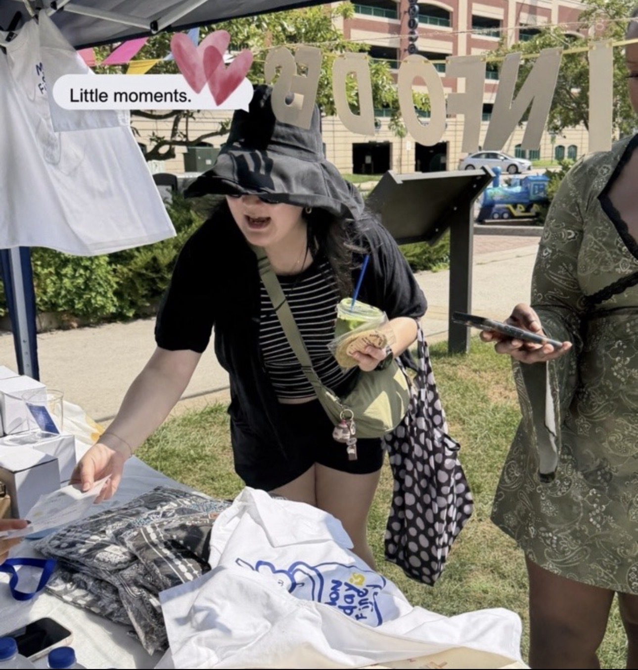 Woman with black hat and striped shirt reaching for items at outdoor market stall, with another person nearby holding a phone.