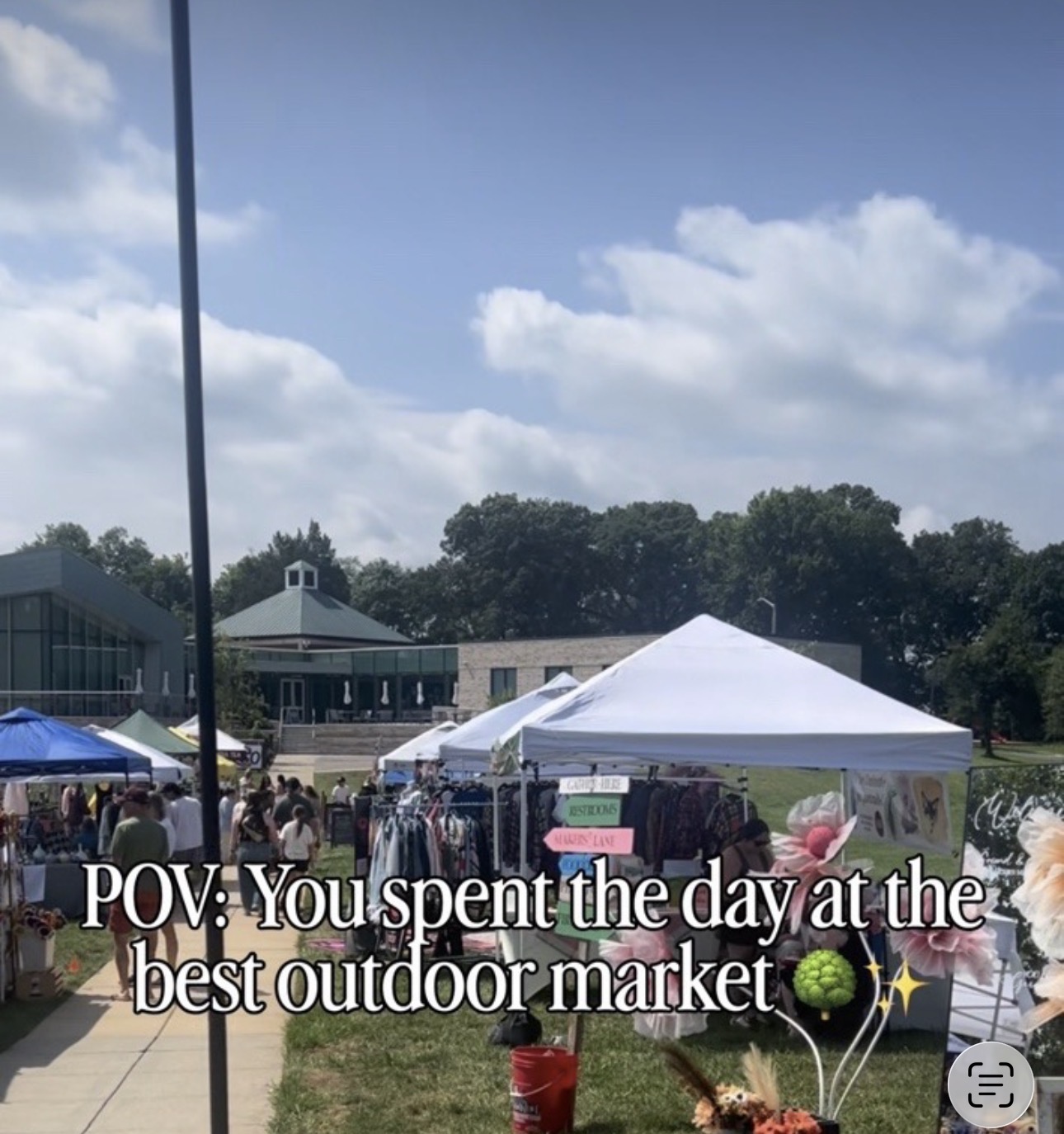 Outdoor market with tents, people, and trees under a partly cloudy sky.