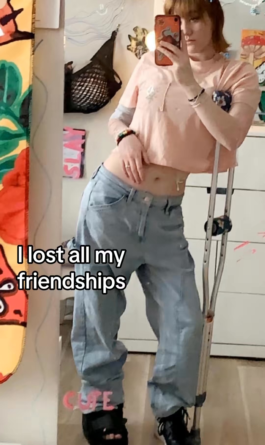 Person taking mirror selfie with crutches, wearing a pink top and baggy jeans, in a room with colorful decor.