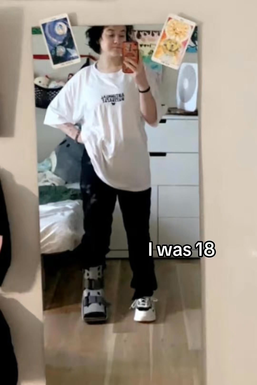 Person taking a mirror selfie in a room with posters on the wall, wearing a white t-shirt and black pants, with text