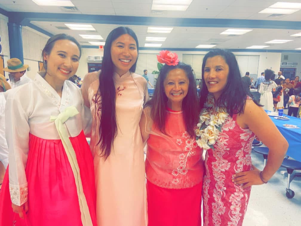 Four women smiling indoors, one wearing a pink dress with a flower headband, others in traditional and floral attire, in a decorated room.