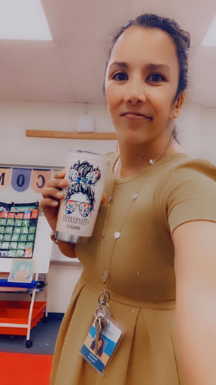 Woman holding a decorated cup in a store, smiling, wearing a beige dress and a lanyard with an ID badge.