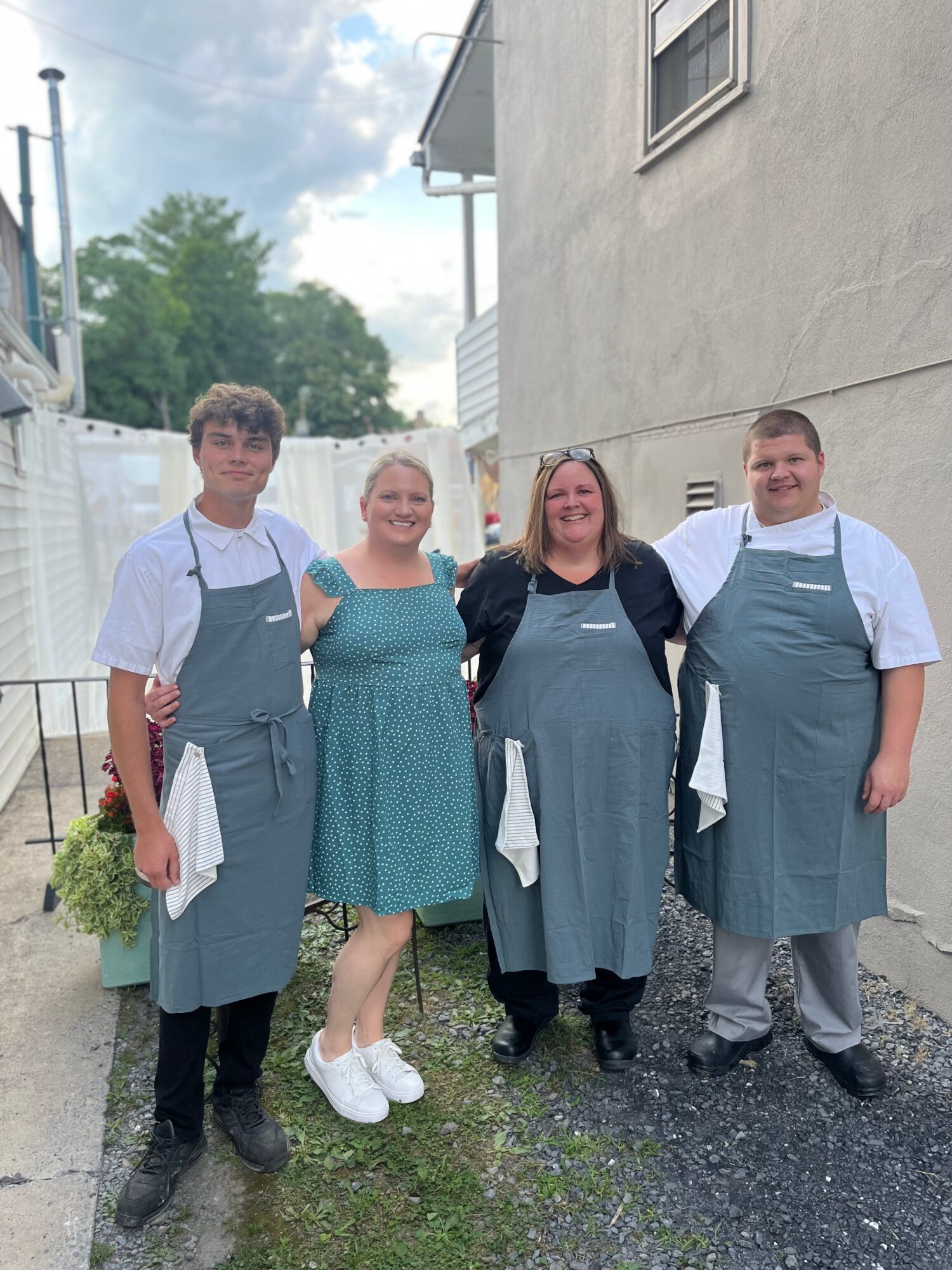 Four people standing outdoors, two women and two men, smiling, wearing aprons, in front of a building with a white wall.