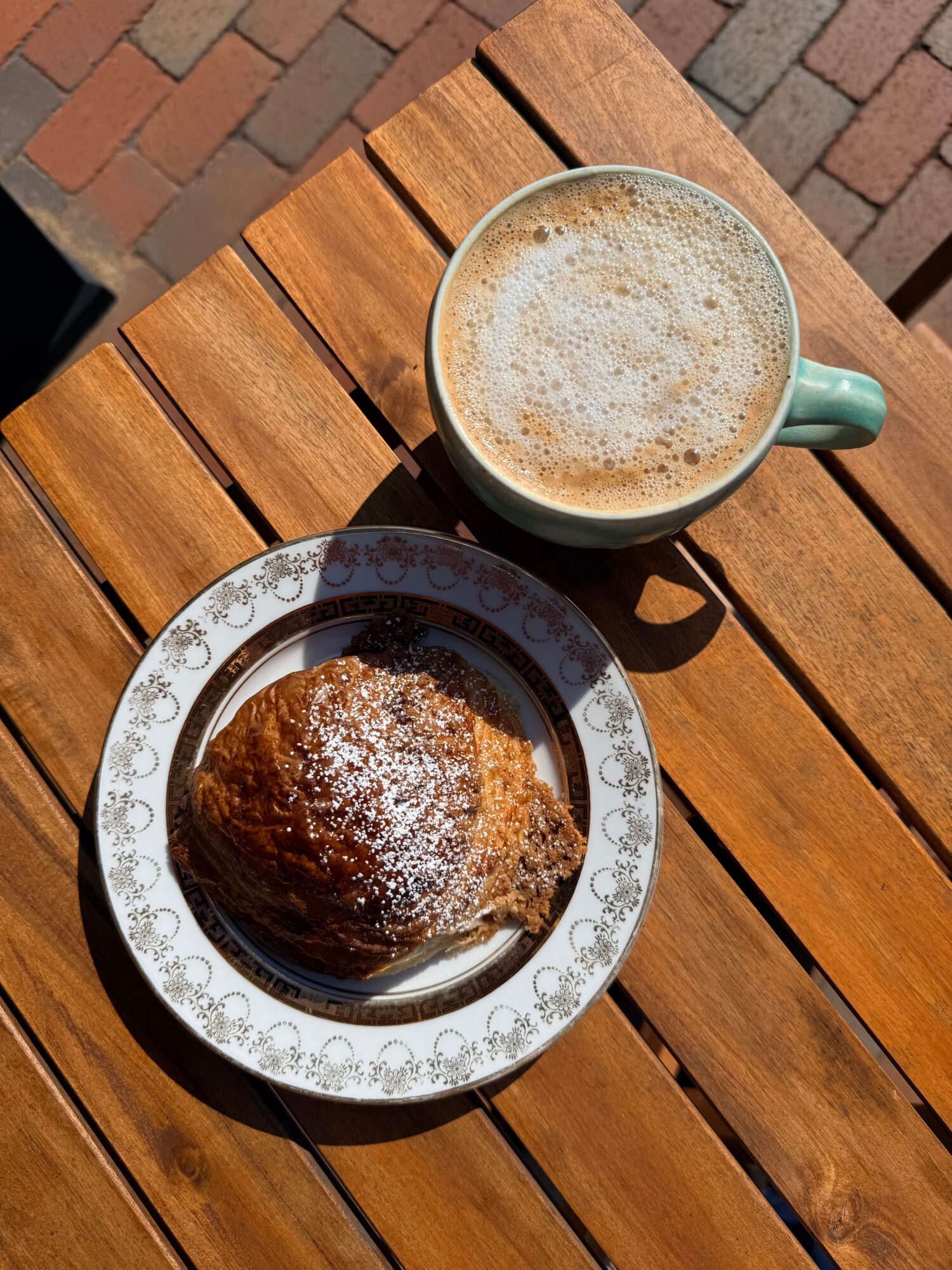 Cup of frothy coffee and a pastry dusted with powdered sugar on a decorative plate, on a wooden table.