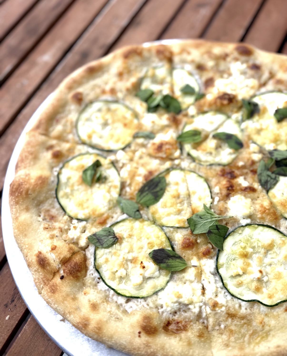 Pizza with cheese, zucchini slices, and basil leaves on a white plate, on a wooden surface.