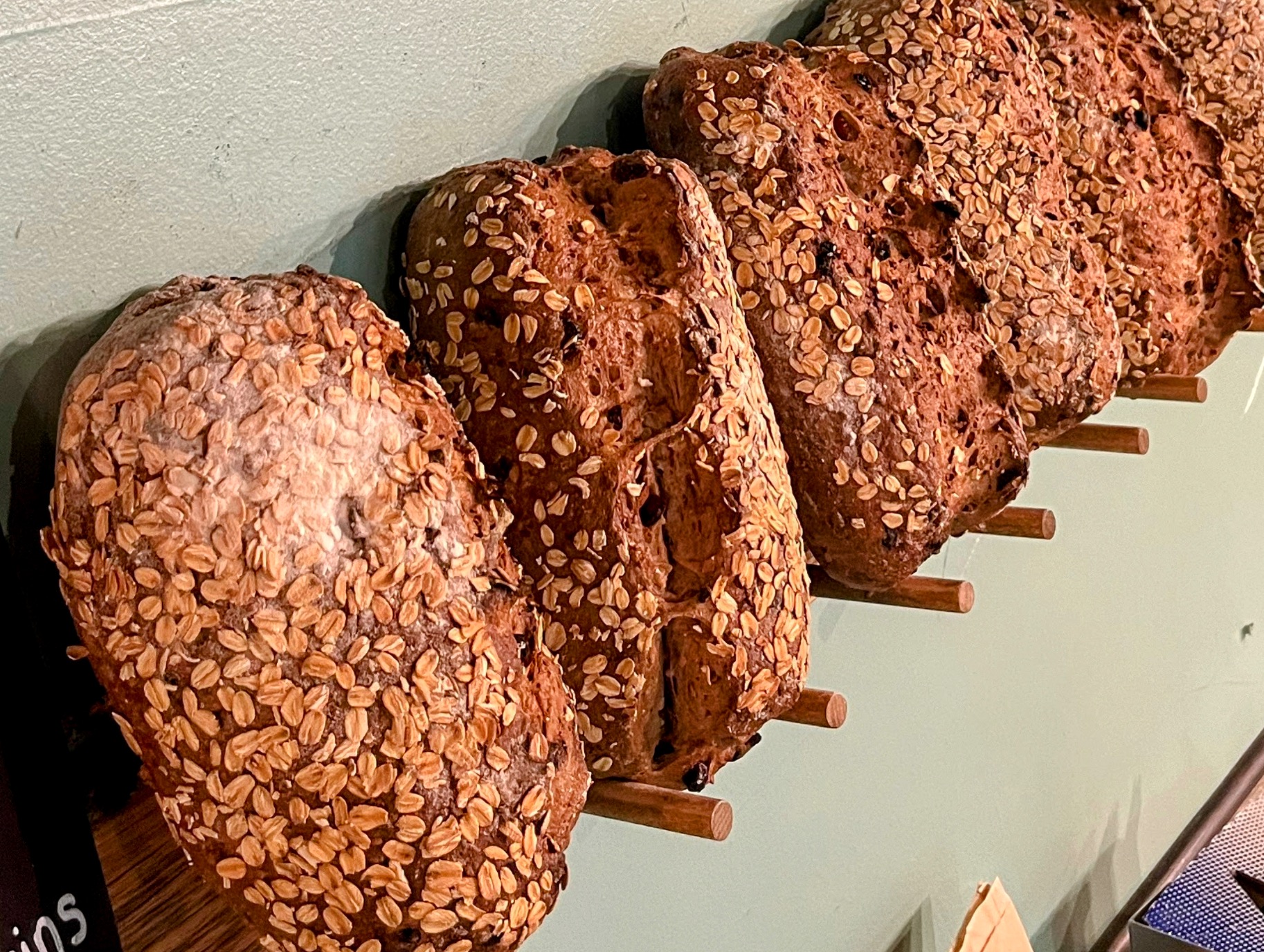 Five large loaves of bread with seeds on top, lined up on a wooden rack against a light-colored wall.