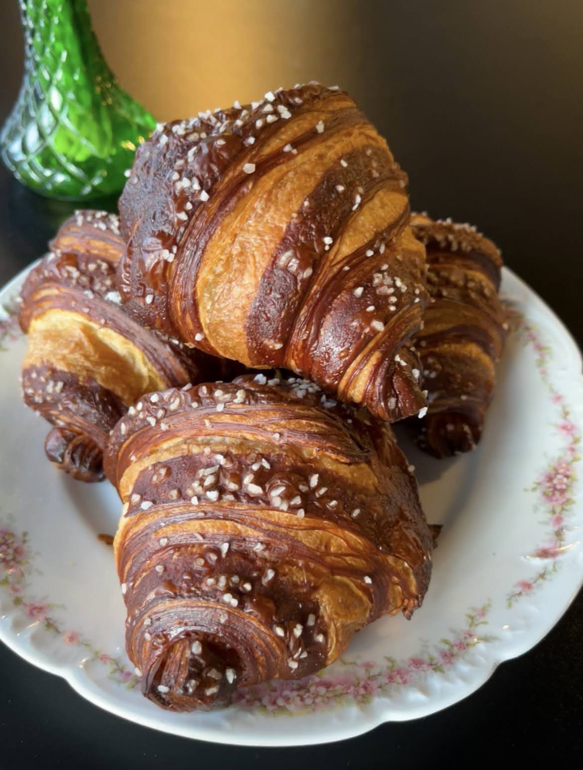 Three croissants with sugar crystals on a floral-patterned plate, with a green bottle in the background.