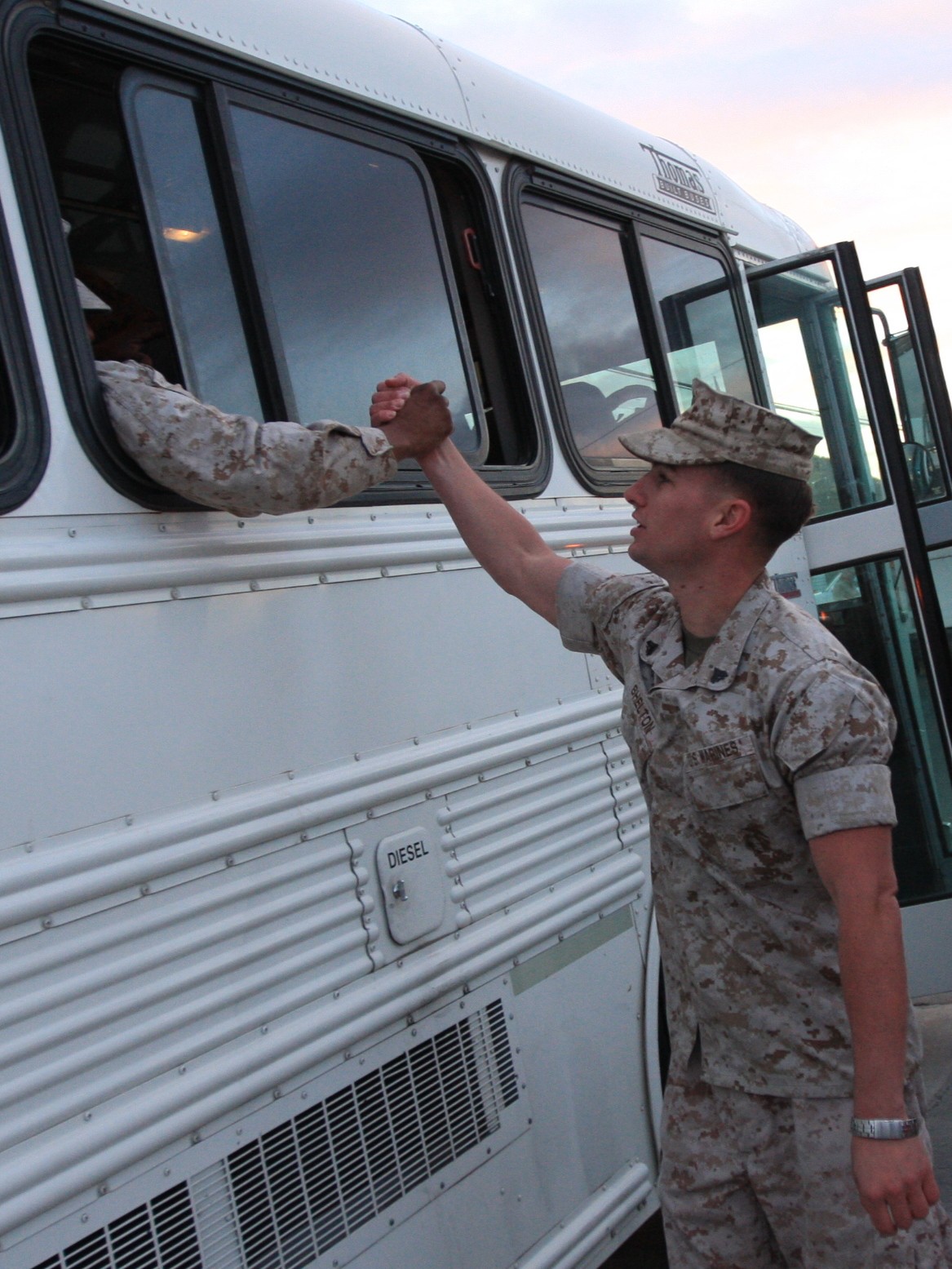 Soldier in uniform shakes hands with person inside bus through window, bus door open, sky in background.