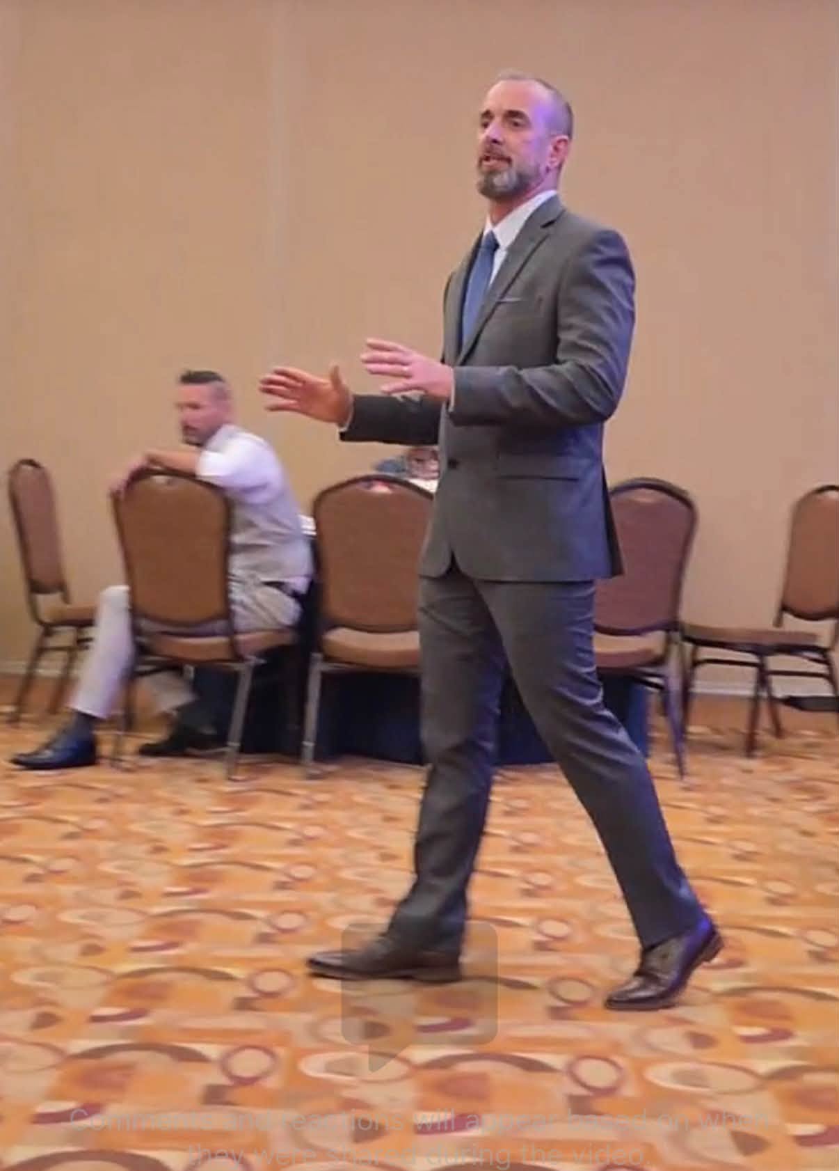 Man in a gray suit standing and gesturing, with chairs and a person sitting in the background, in a room with patterned carpet.
