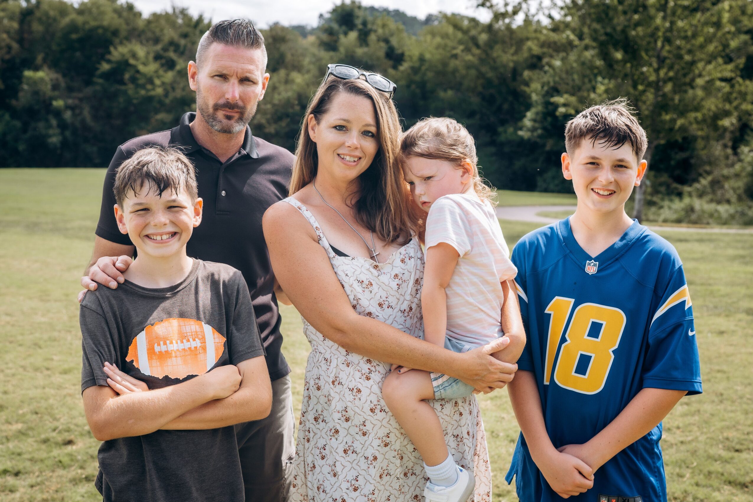 Family of five outdoors, two children, two adults, smiling, standing on grass with trees in background.