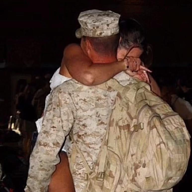 Two soldiers embrace, one wearing a camouflage uniform and the other with a backpack, in a dark setting.
