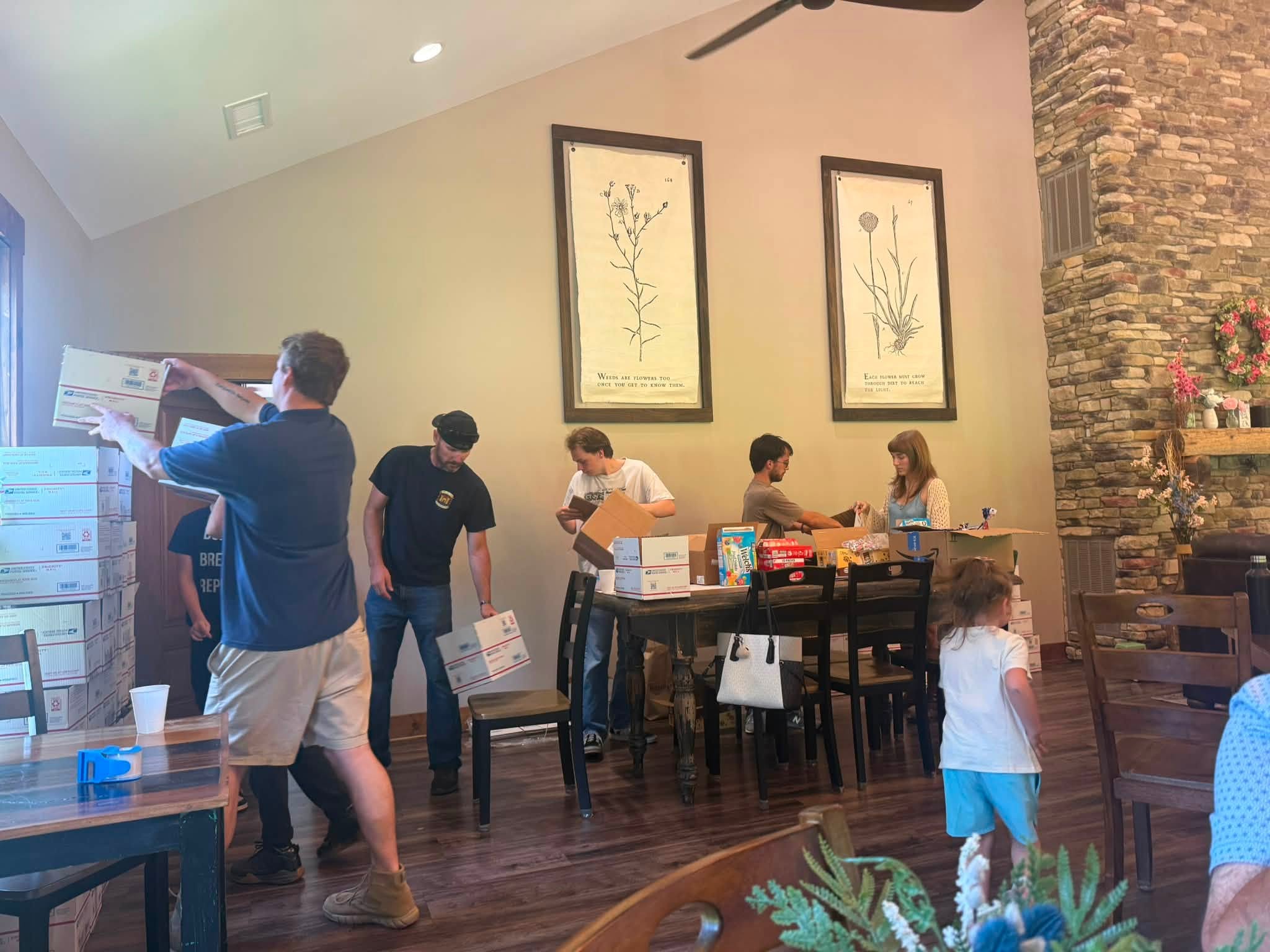 People gather around a dining table in a room with wall art and a stone fireplace, some standing and some seated.