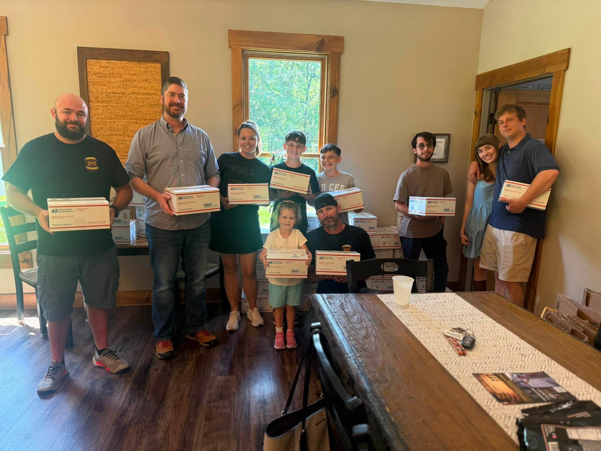 Group of ten people, including children, holding boxes in a room with wooden floors and windows, smiling at the camera.