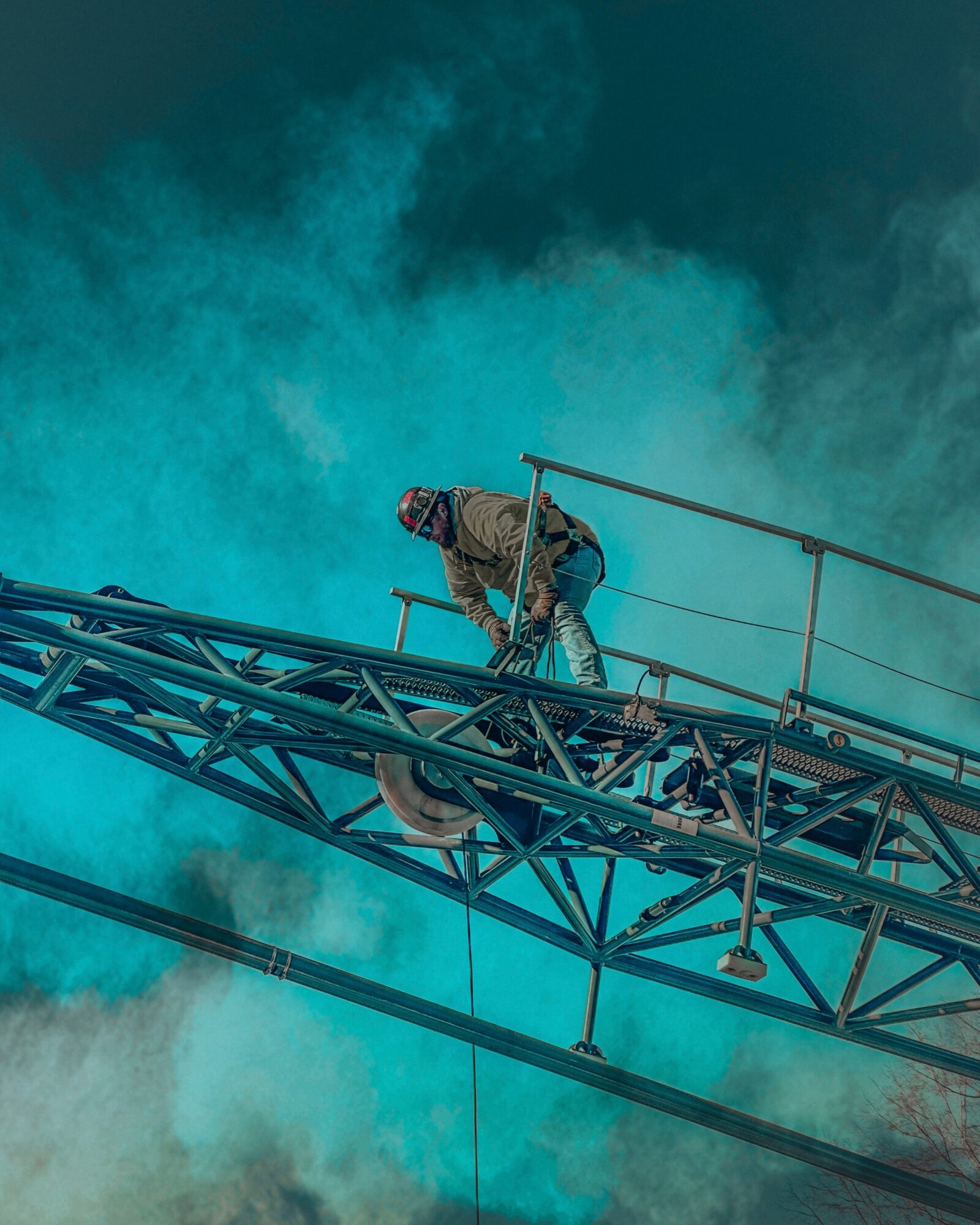 Worker in safety gear on a tall metal tower against a cloudy sky, performing maintenance or inspection.