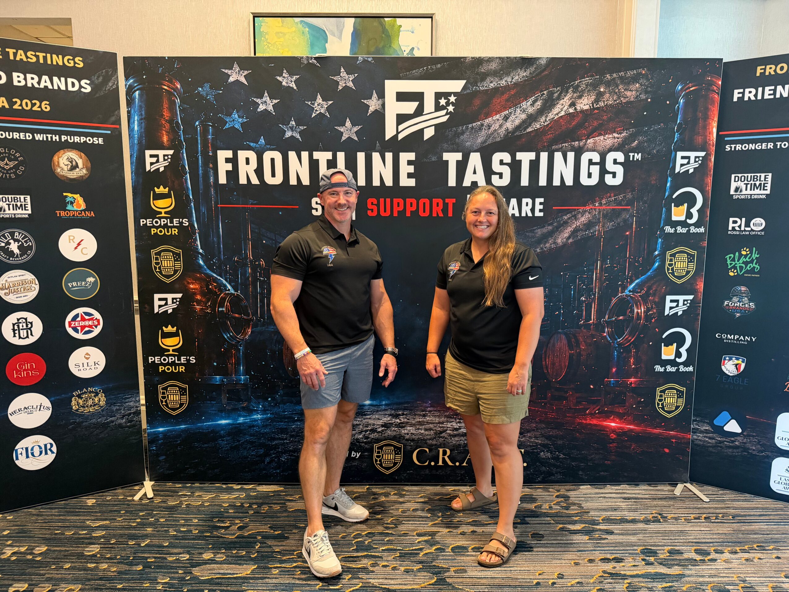Two people stand in front of a large banner with logos and text, smiling at the camera.