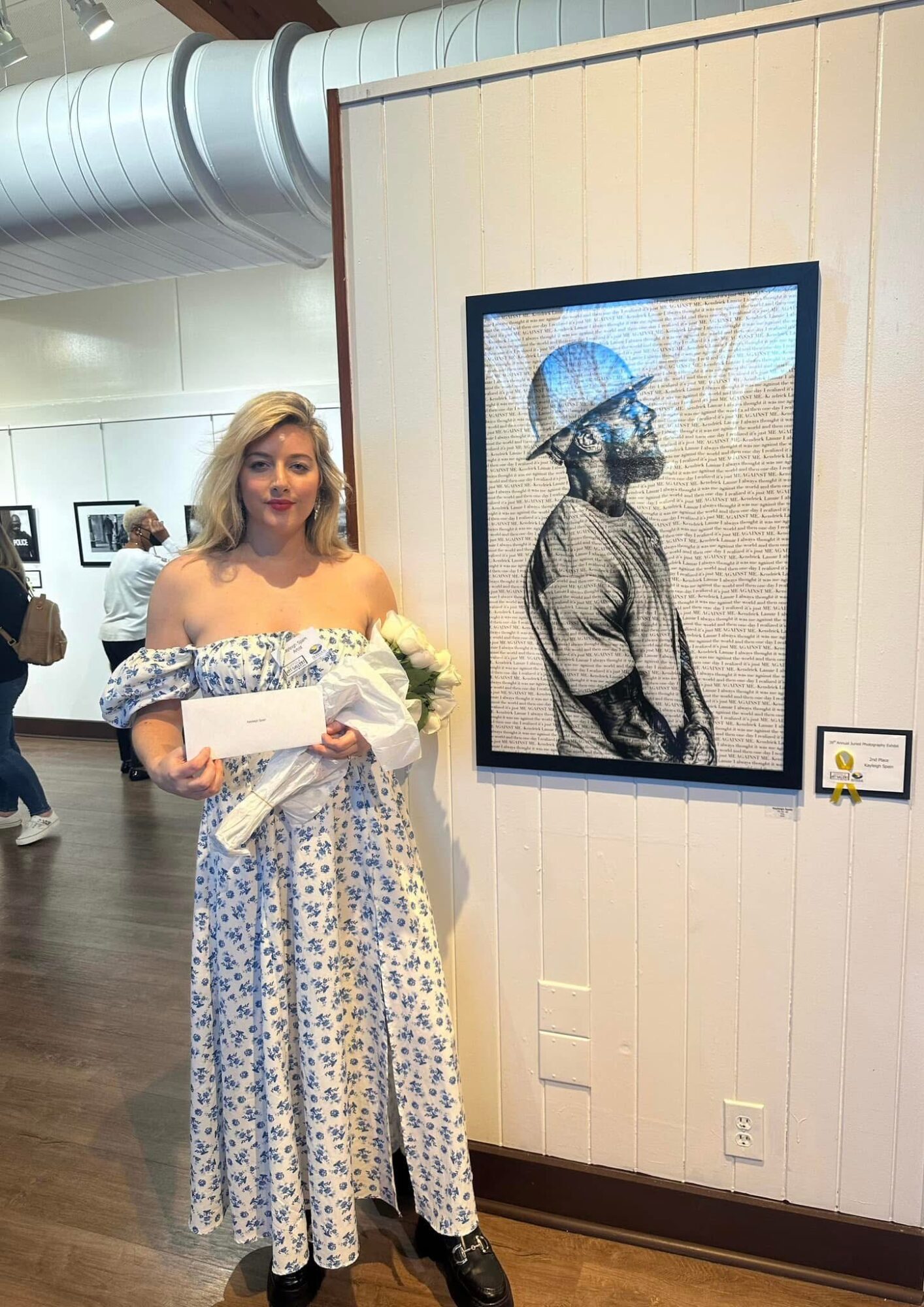 Young woman in a floral dress holding a bouquet and envelope, standing next to a framed artwork of a person wearing a helmet, in an art gallery.