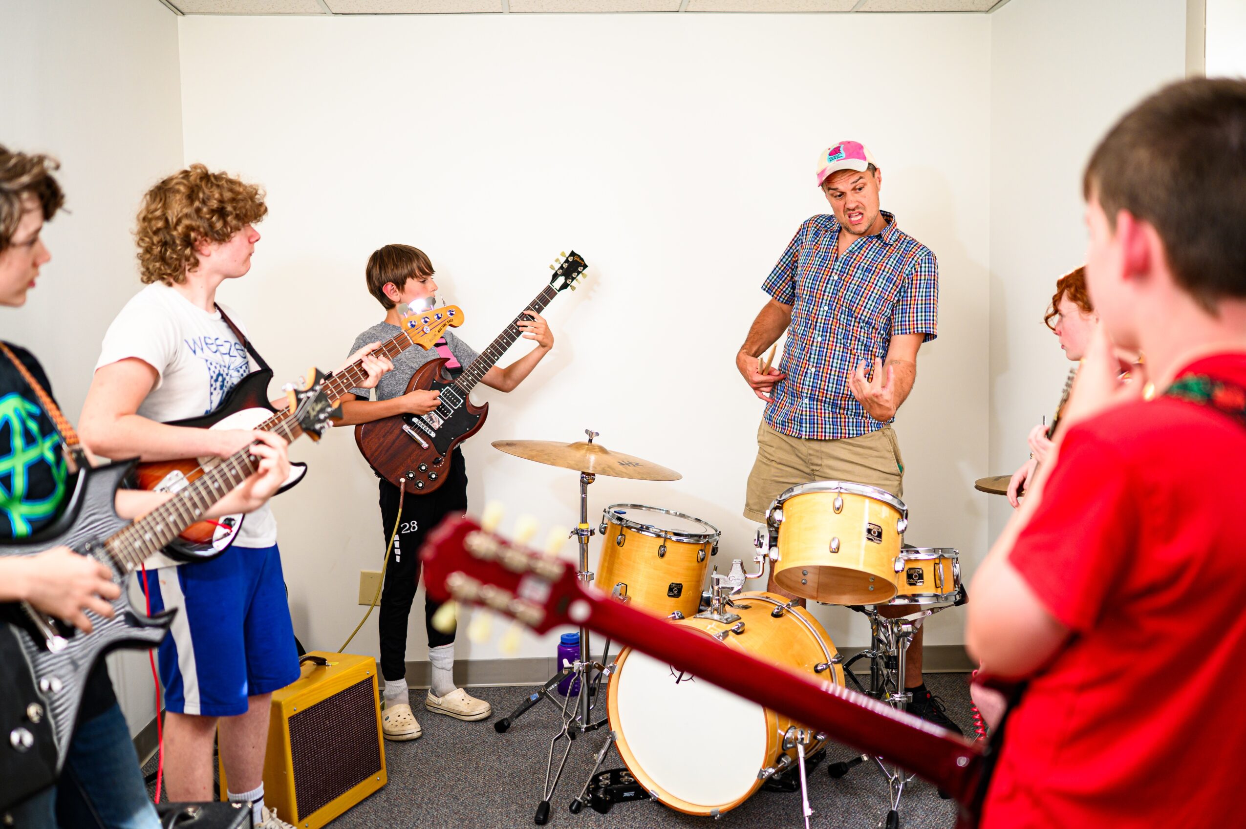 Group of children playing guitars and drums in a music room, with a person leading the session, all engaged and focused.
