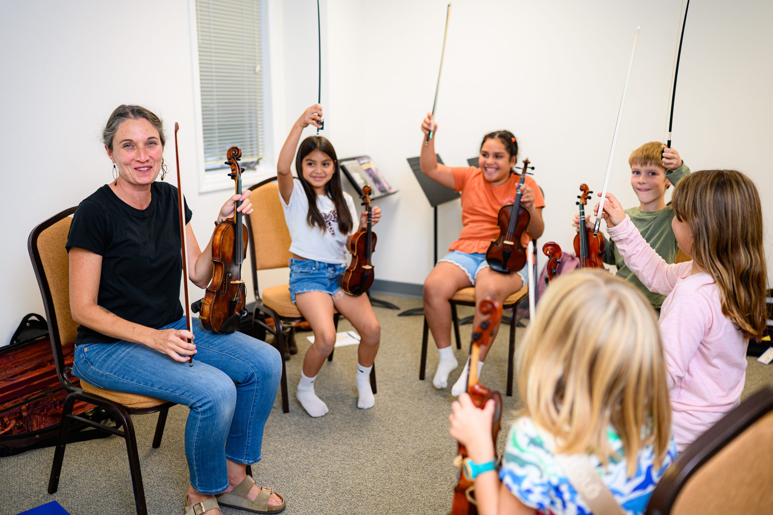 Music teacher with children holding violins in a classroom, smiling and raising bows, group sitting on chairs and floor.