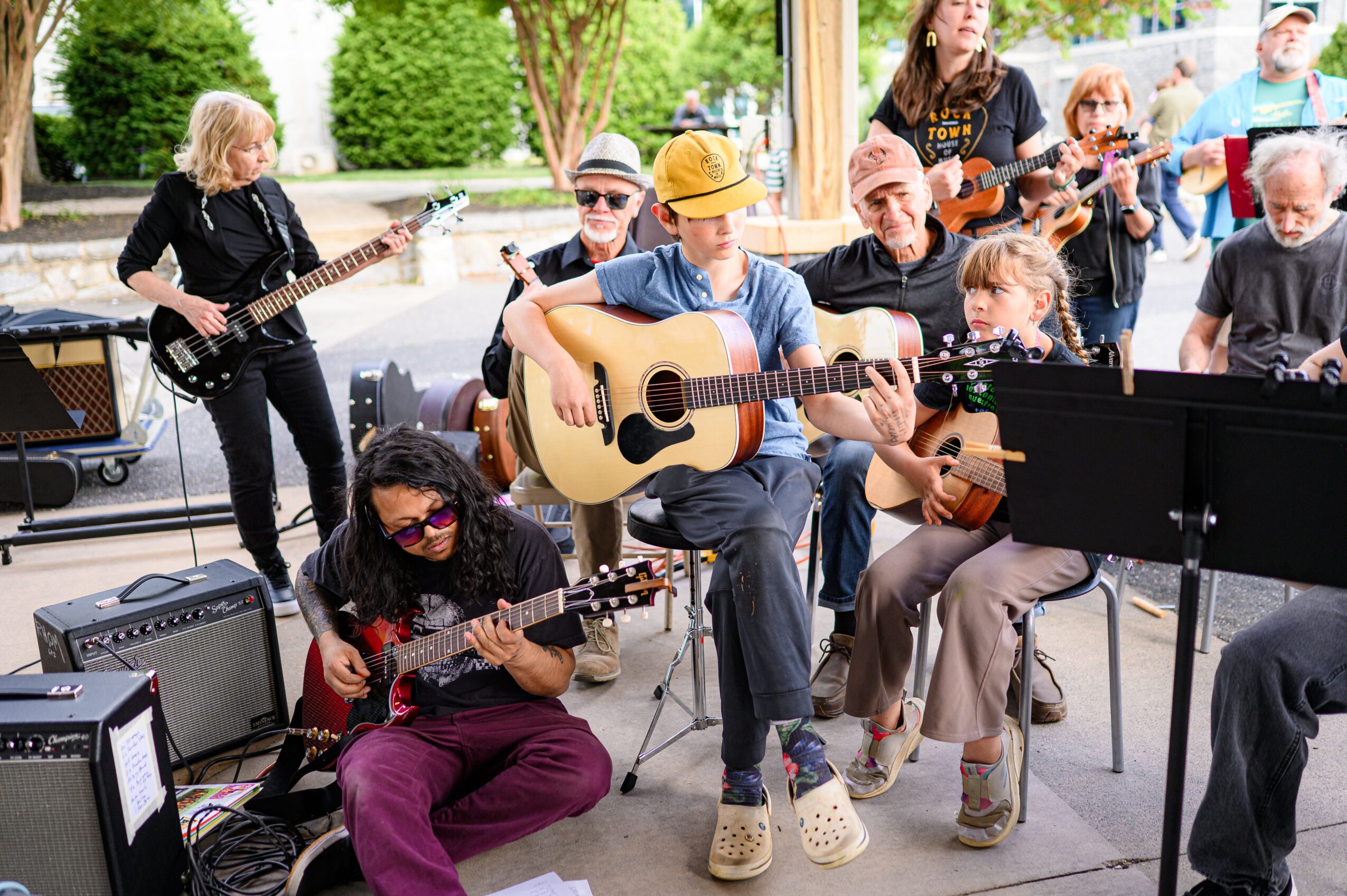 Group of children and adults playing guitars and singing outdoors under a pavilion, with trees and a fence in the background.