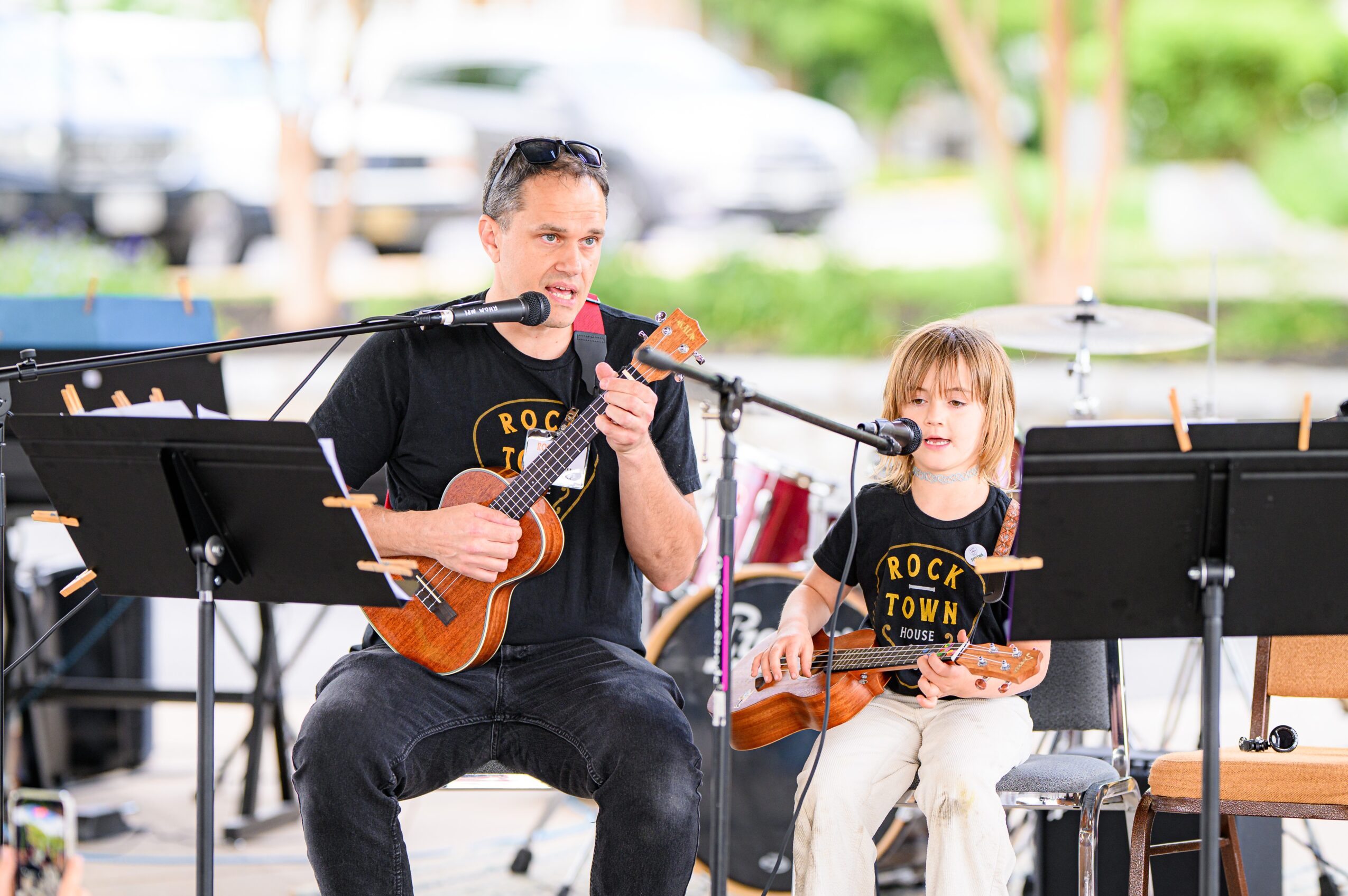 Man and girl playing ukuleles outdoors, singing into microphones, with music stands around them.