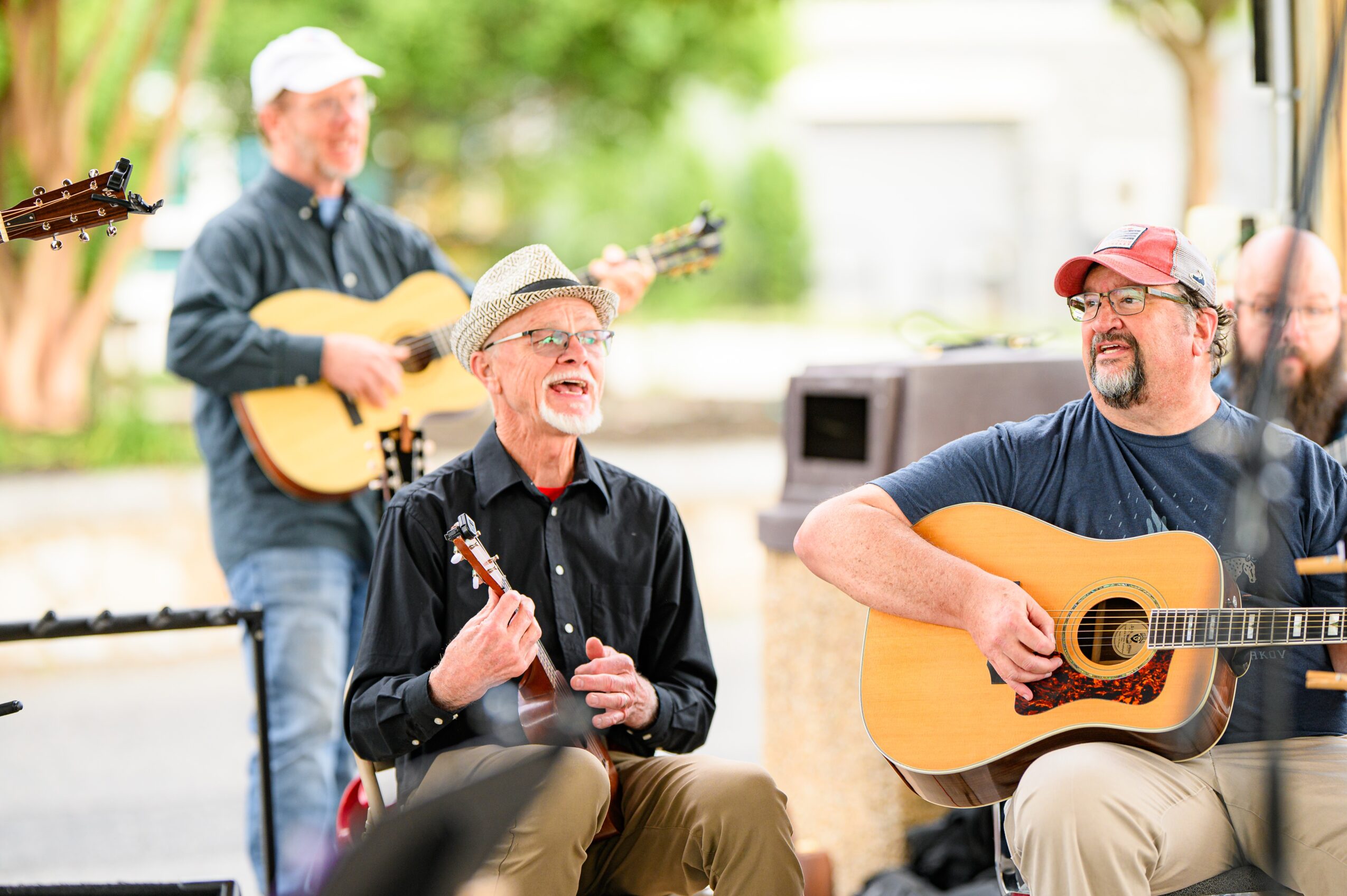 Three men playing guitars outdoors, one singing, with trees and a building in the background.