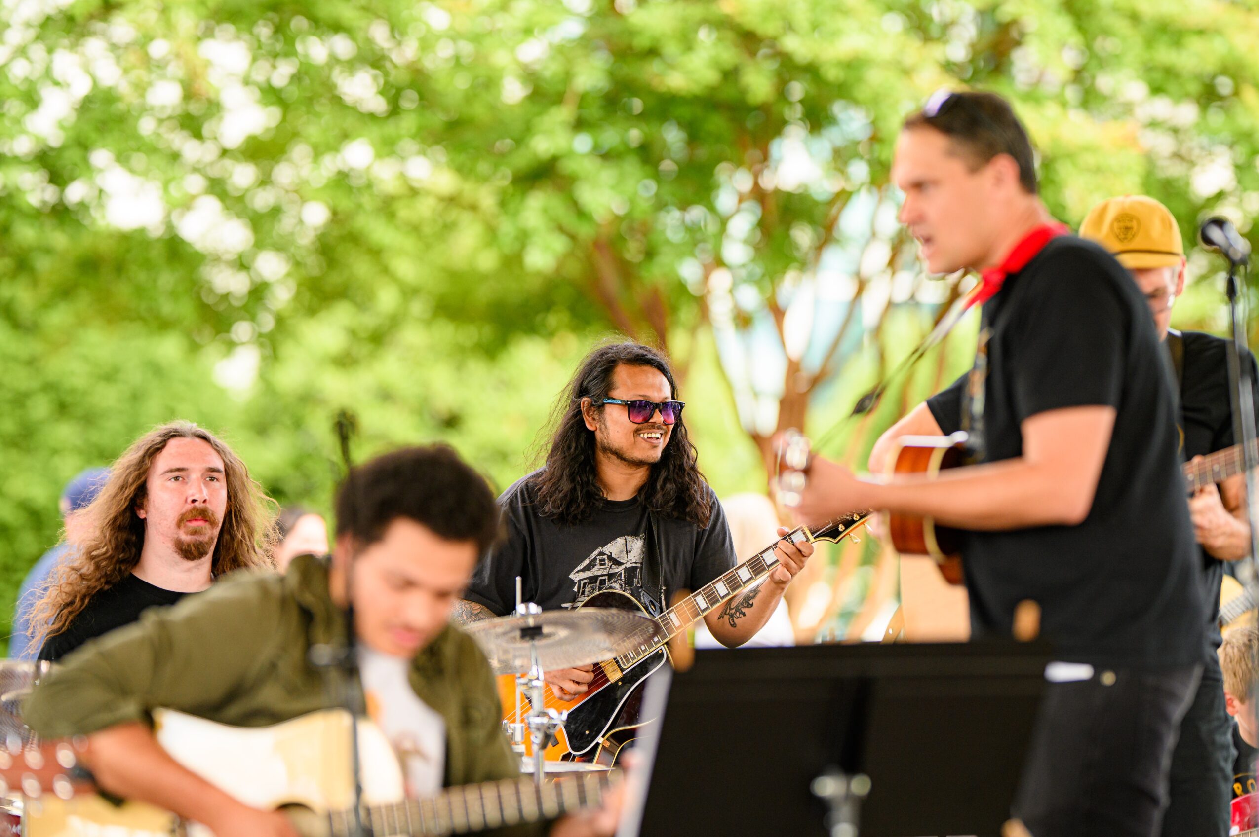 Group of people outdoors, one playing guitar, others listening, with trees and sunlight in background.