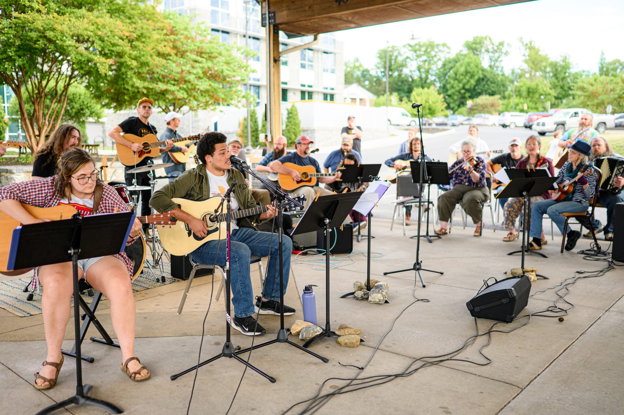 Group of people playing guitars and singing outdoors under a shelter, with trees and parked cars in the background.