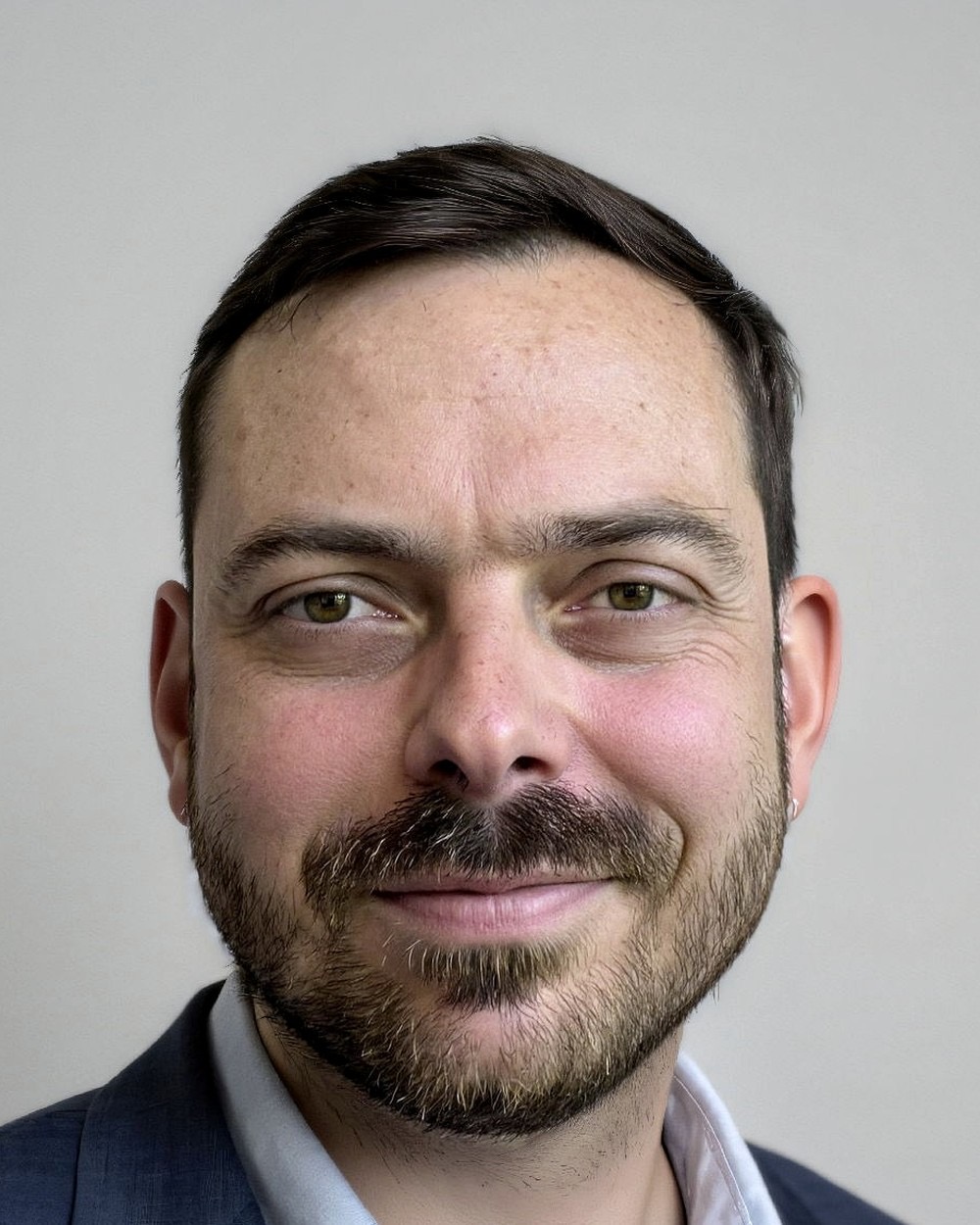 Close-up portrait of a man with short dark hair, beard, and mustache, smiling slightly, wearing a dark suit and light shirt, neutral background.